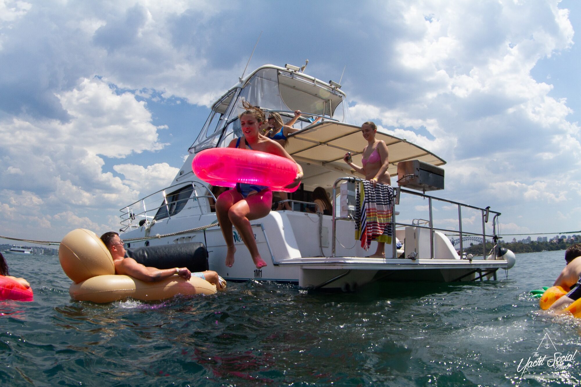 A small group of people are enjoying a sunny day on the water. One person is jumping off a luxurious boat from The Yacht Social Club with a pink inflatable ring, while others float nearby on various inflatable rafts. The background shows a partly cloudy sky and more water.