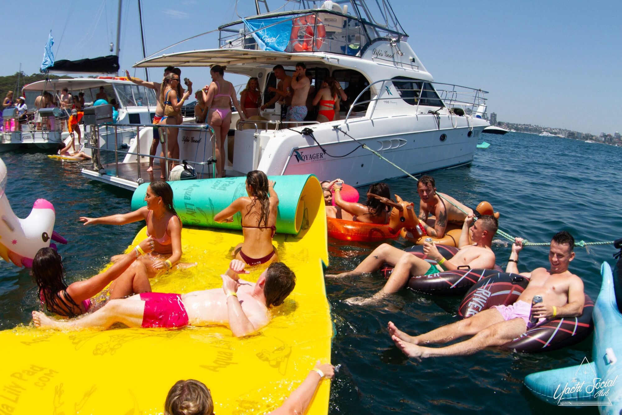 People enjoying a sunny day on a lake, floating on various inflatable rafts and lounging on a large yellow mat. A yacht from The Yacht Social Club Sydney Boat Hire is anchored nearby with more people on board. The atmosphere is fun and relaxed.