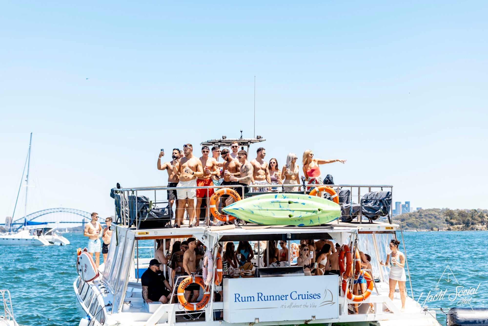 A large group of people in swimsuits stands on the upper deck of a boat named "Rum Runner Cruises," enjoying the sunny weather. Some passengers are posing and waving, with a city skyline and bridge visible in the background over the blue water. Experience unforgettable moments with The Yacht Social Club's Boat Parties Sydney.