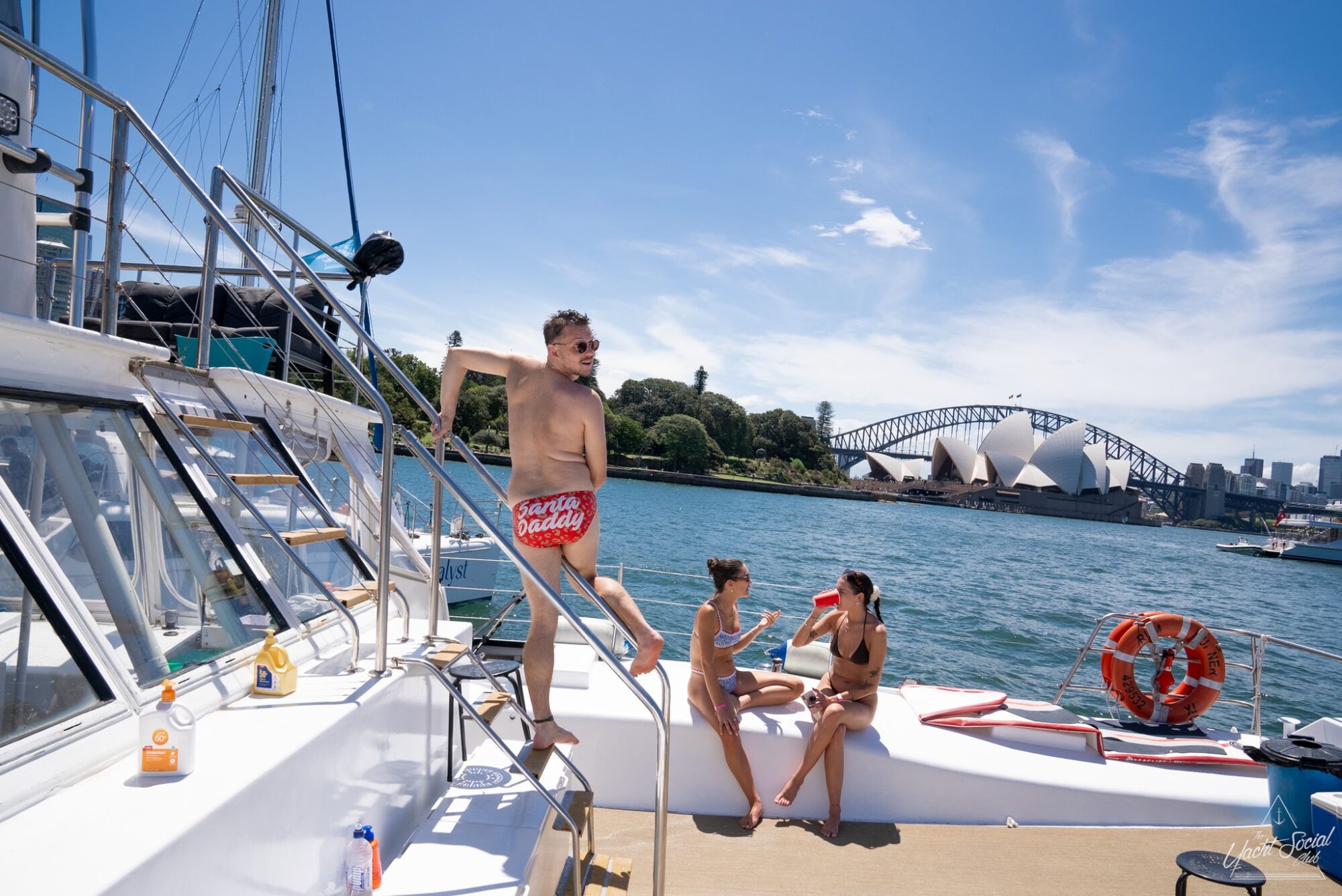 A man stands on the steps of a boat wearing red swim trunks that say "sugar daddy." Nearby, two people in swimsuits lounge on the deck. The background features a clear blue sky, water, and the Sydney Opera House as well as the Harbour Bridge—a perfect setting for The Yacht Social Club's luxury yacht rentals in Sydney.