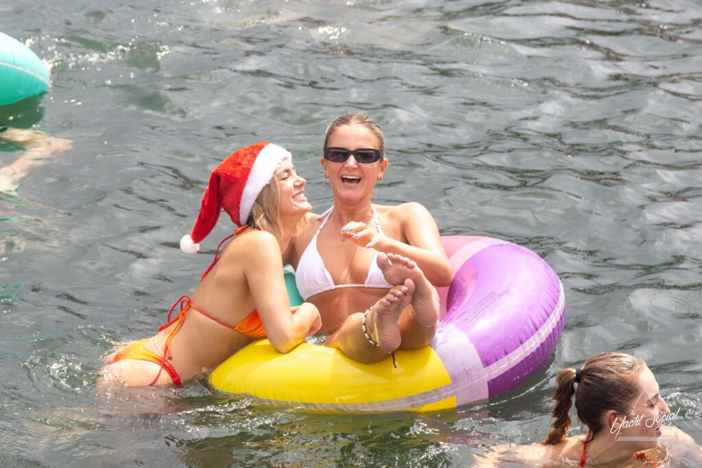 Two women enjoying the water; one wears a Santa hat and a red bikini, while the other wears sunglasses and a white bikini, lounging on a colorful inflatable ring. They are laughing and have their arms around each other, with The Yacht Social Club Event Boat Charters creating the perfect backdrop.