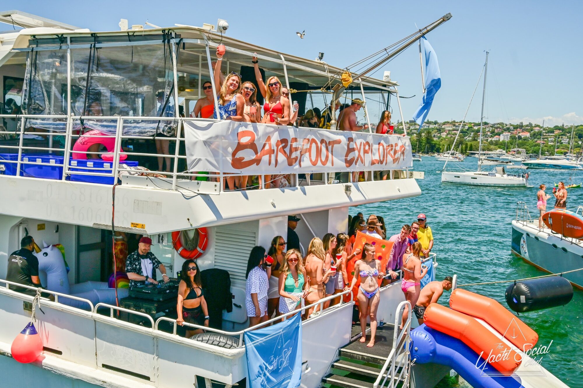 A lively party is taking place on a double-decker boat named "Barefoot and Boujee" docked near other boats. People in swimwear are on both decks, some waving at the camera and others near the water. Hosted by The Yacht Social Club Sydney Boat Hire, a large unicorn float is tied to the side of the boat.