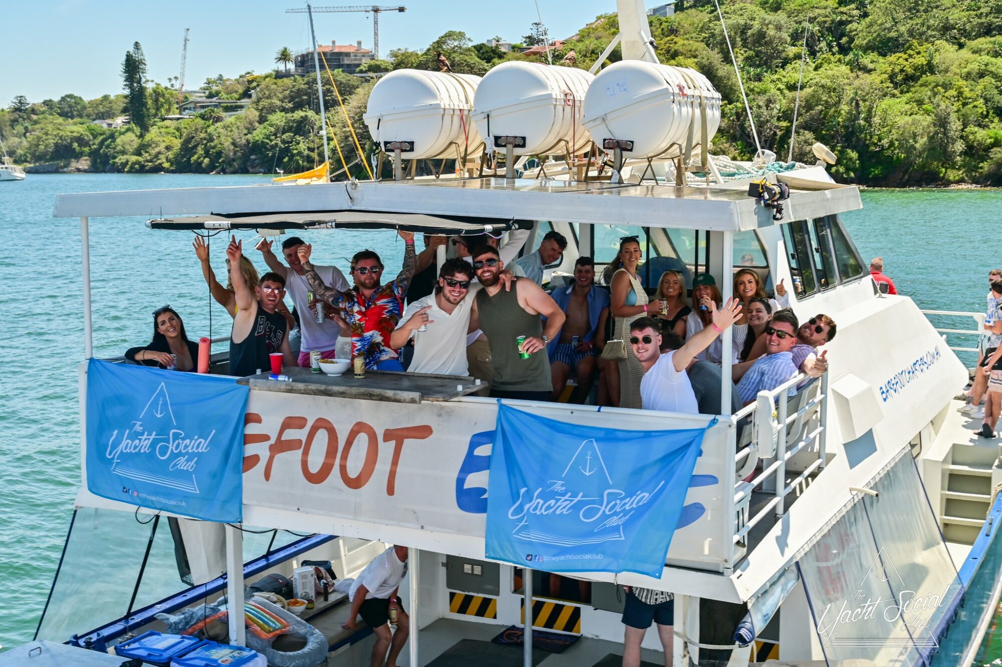 A group of people smiling and waving on a boat named "Seafood," docked in a scenic area with greenery in the background. Many are wearing sunglasses and summer attire. Banners on the boat read "The Yacht Social Club," perfect for those interested in Sydney Harbour Boat Hire The Yacht Social Club.