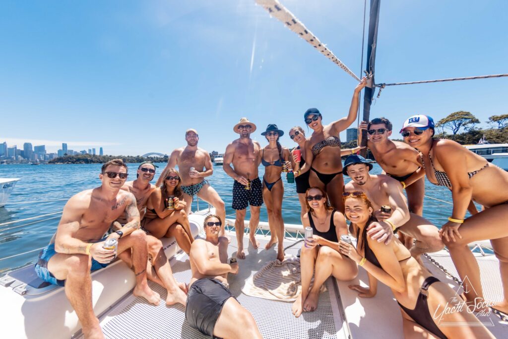 A group of people in swimwear enjoy a sunny day on a boat from The Yacht Social Club Sydney Boat Hire. Some are standing and posing, while others sit or recline. They smile and hold drinks, with a scenic view of water and a city skyline in the background.