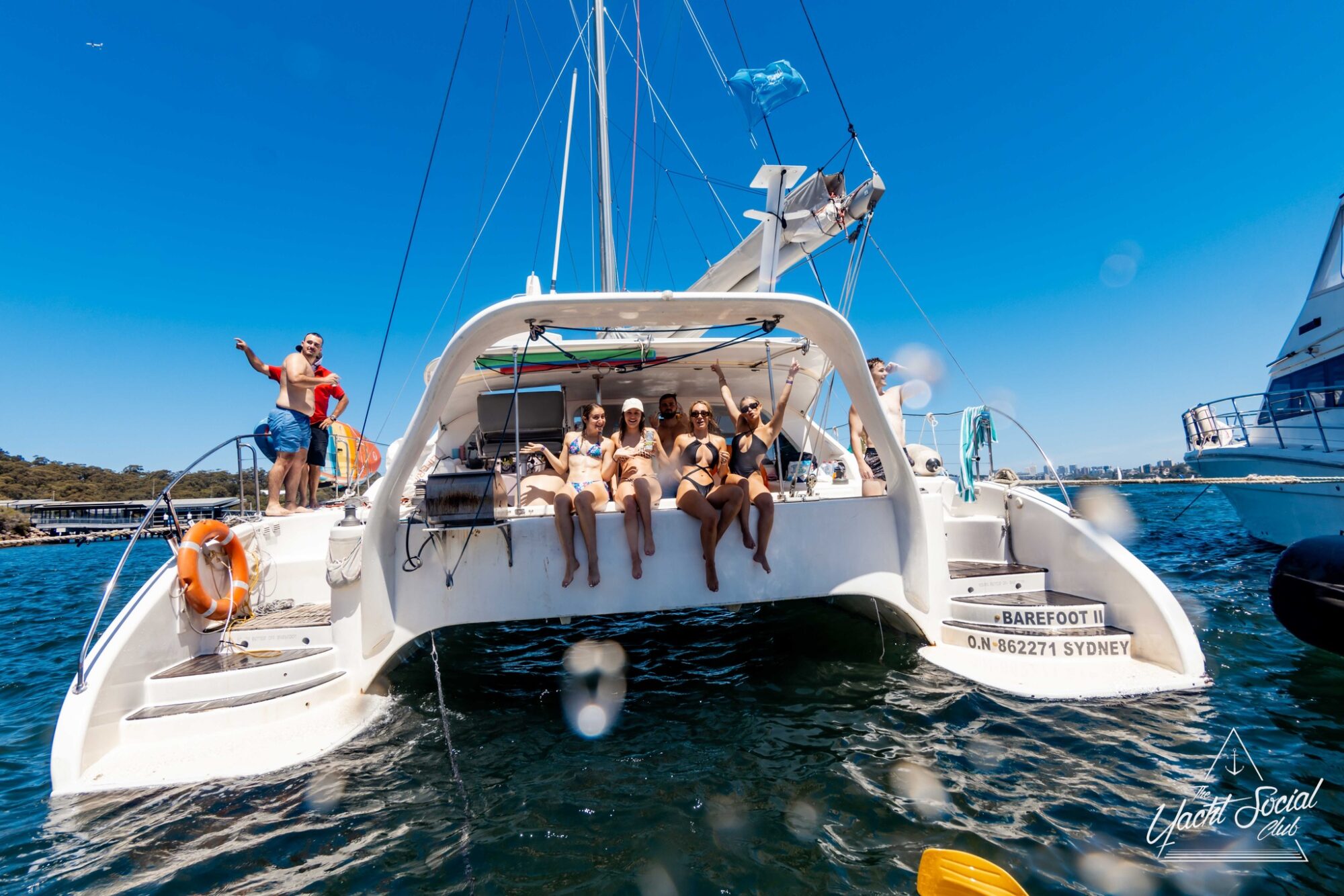 A group of people sitting and relaxing on the stern of a white catamaran named "Barefoot" with the registration "ON 862711 SYDNEY." The Yacht Social Club adds a touch of elegance to this sunny, scenic setting. Clear blue skies frame the boat, with another vessel visible in the background.