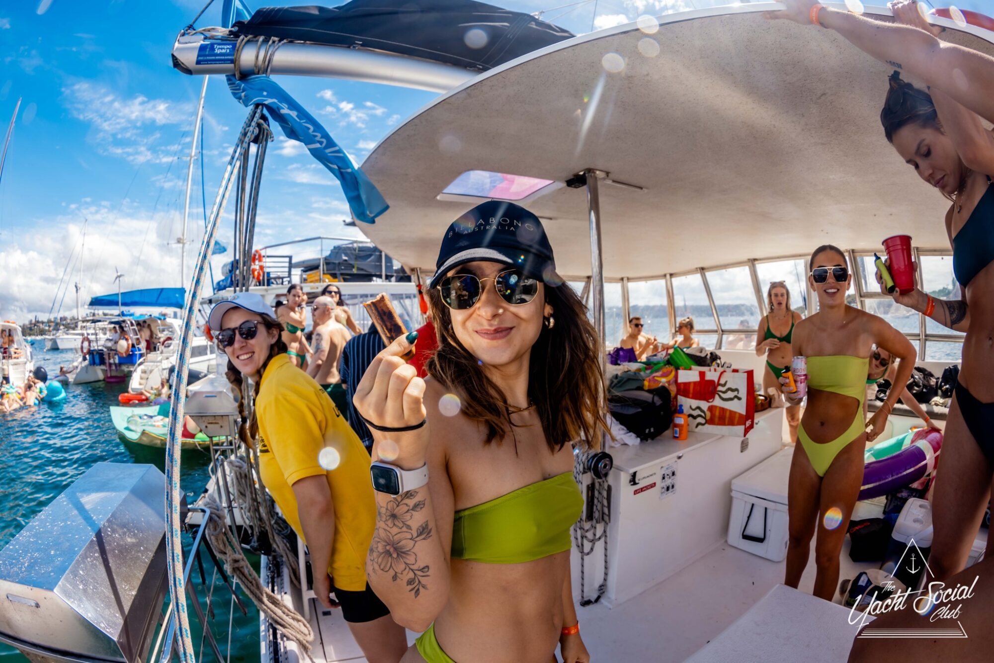 A group of people enjoying a vibrant boat party under a sunny sky. The central figure, a woman in a green bikini and sunglasses, smiles while holding a snack stick. Other attendees in swimwear interact in the background. Hosted by The Yacht Social Club Event Boat Charters, the atmosphere is festive and lively.