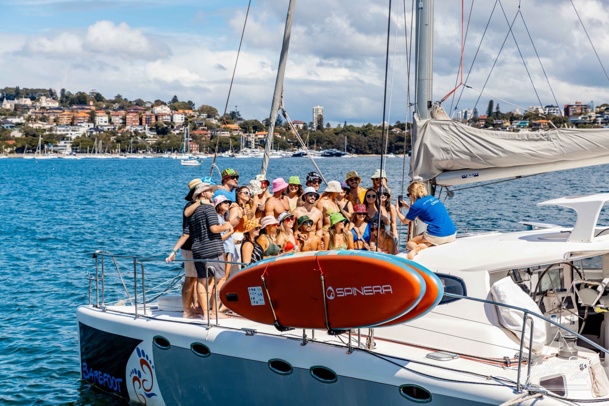 A group of people stands and sits on the deck of a sailboat during The Yacht Social Club Event. The sailboat is on a body of water with a shoreline and buildings visible in the background. An orange Spinera paddleboard is secured to the boat, adding to the lively atmosphere.