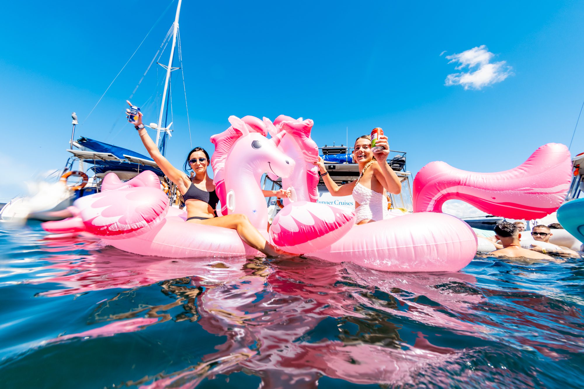 Two people sit on large pink inflatable flamingos, smiling and holding drinks, while enjoying the water near a luxury yacht. The sky is clear and blue with other people and inflatables around them. Experience the ultimate day out with Sydney Harbour Boat Hire by The Yacht Social Club.