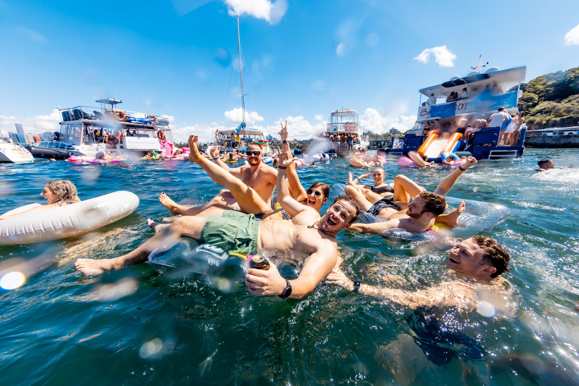 A group of people having fun in the water, floating on inflatable tubes and holding drinks, with boats from The Yacht Social Club Sydney Boat Hire and a clear blue sky in the background. Some are smiling and waving at the camera, with lush green trees visible in the distance.