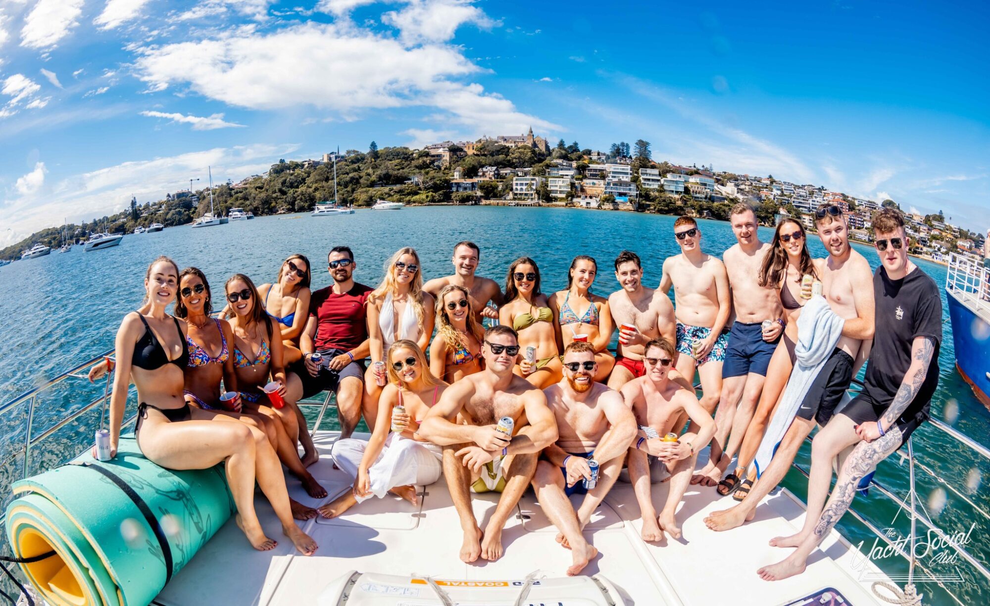 A large group of adults in swimwear pose for a photo on a boat under a sunny sky. The backdrop features a body of water, green shoreline, and houses on a hillside. Everyone is smiling and enjoying the outing with various drinks and relaxed postures at The Yacht Social Club Event Boat Charters.