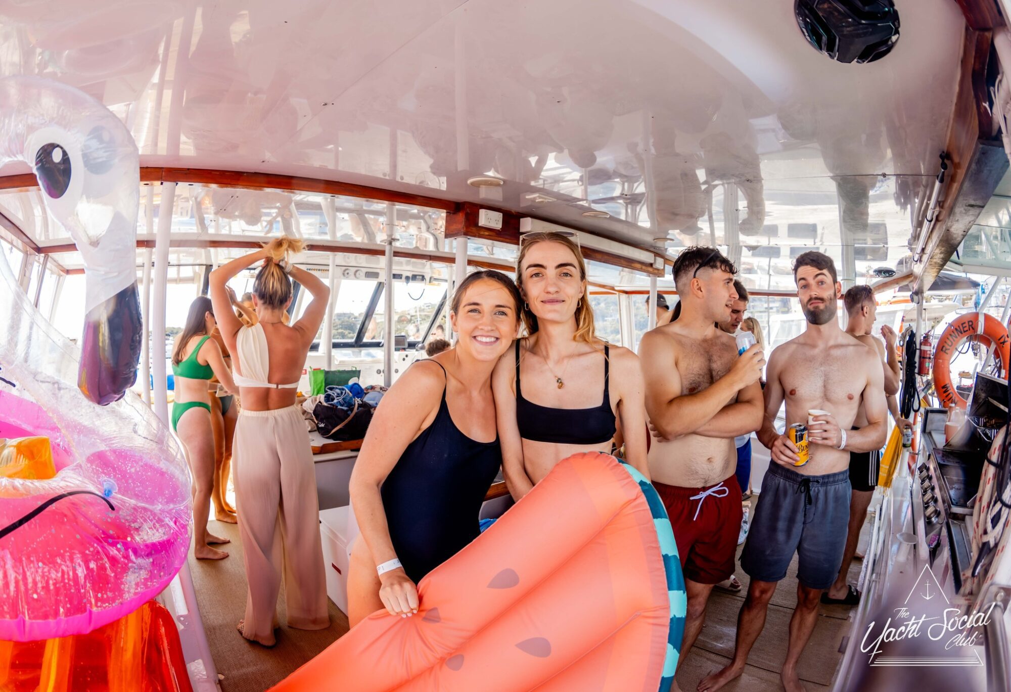 A group of people enjoying a party on a luxury yacht. Two women in the foreground are smiling at the camera and holding a large inflatable watermelon slice. Other partygoers in swimwear are chatting and holding drinks, embodying the spirit of Boat Parties Sydney The Yacht Social Club, with pool toys and life vests visible.