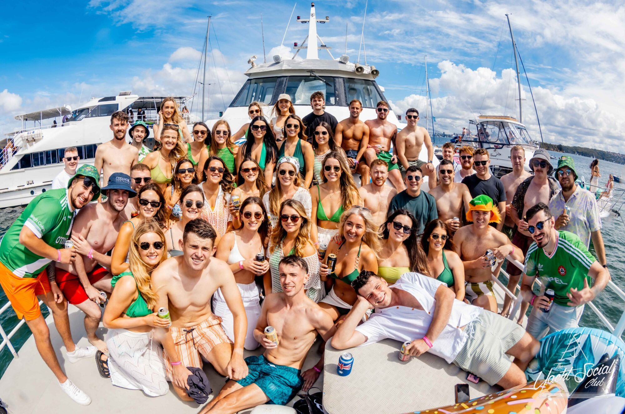 A large group of people in swimwear are gathered on a boat under a sunny sky, enjoying one of the lively Boat Parties Sydney The Yacht Social Club. They are posing for a photo, many smiling and holding drinks, against a backdrop of another boat and water. Some wear sunglasses, and the mood appears festive and fun.