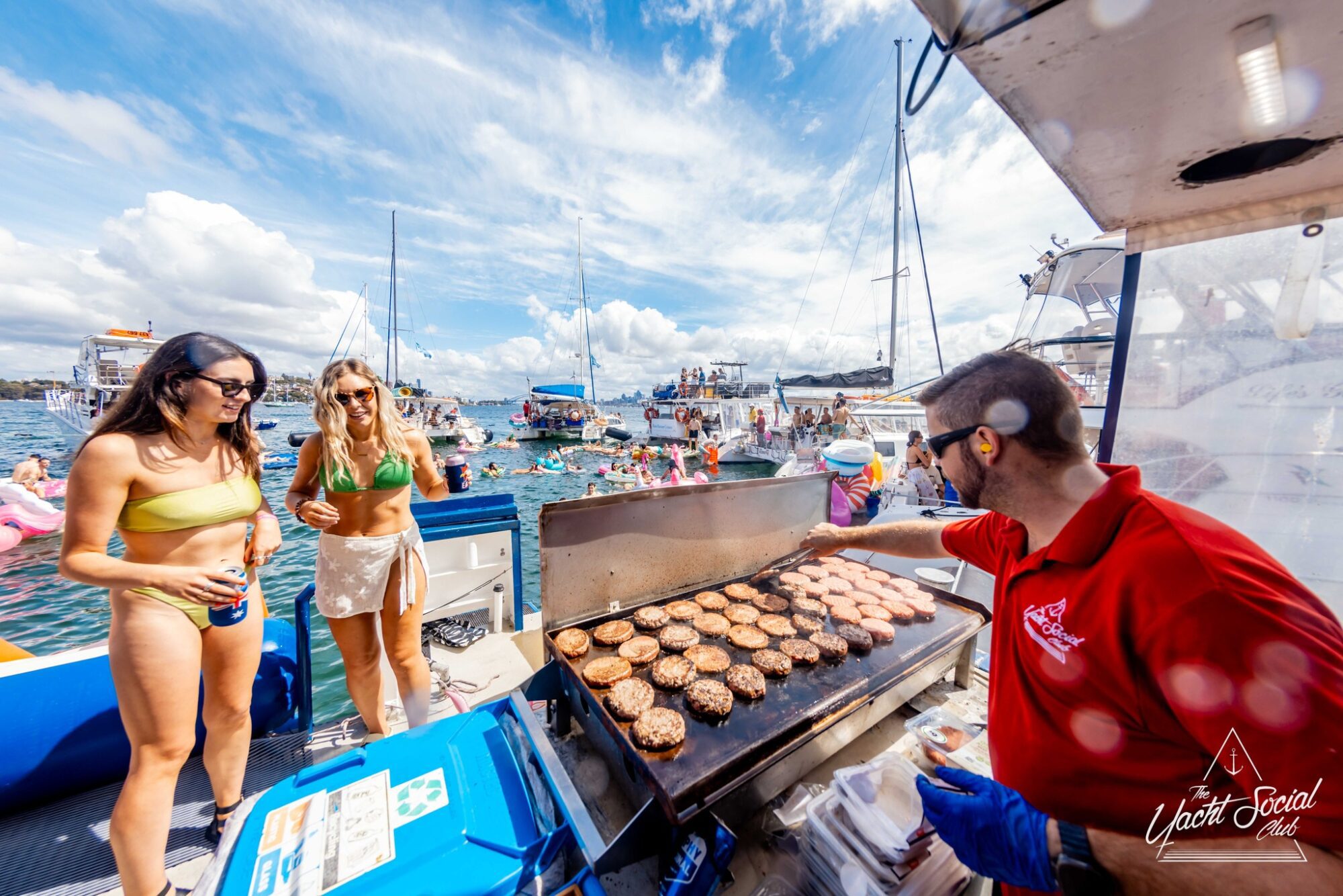 A person in a red shirt is grilling burgers on a yacht from The Yacht Social Club, while two people in swimsuits stand nearby. The background features other yachts and people swimming under a partly cloudy sky. The atmosphere is lively, suggesting a social event or party organized by Sydney Harbour Boat Hire.