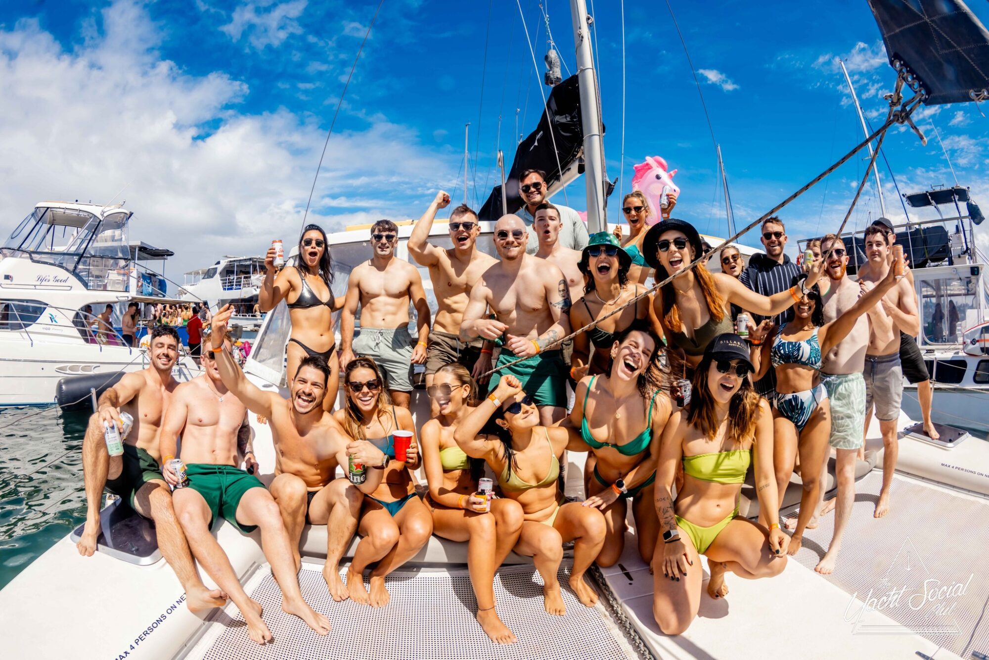 A group of around 20 people, mostly in swimwear, are posing and smiling on a luxury yacht under a partly cloudy sky. They’re holding drinks and appear to be celebrating. Other boats are visible in the background. The scene reflects a lively and festive atmosphere, typical of The Yacht Social Club events.