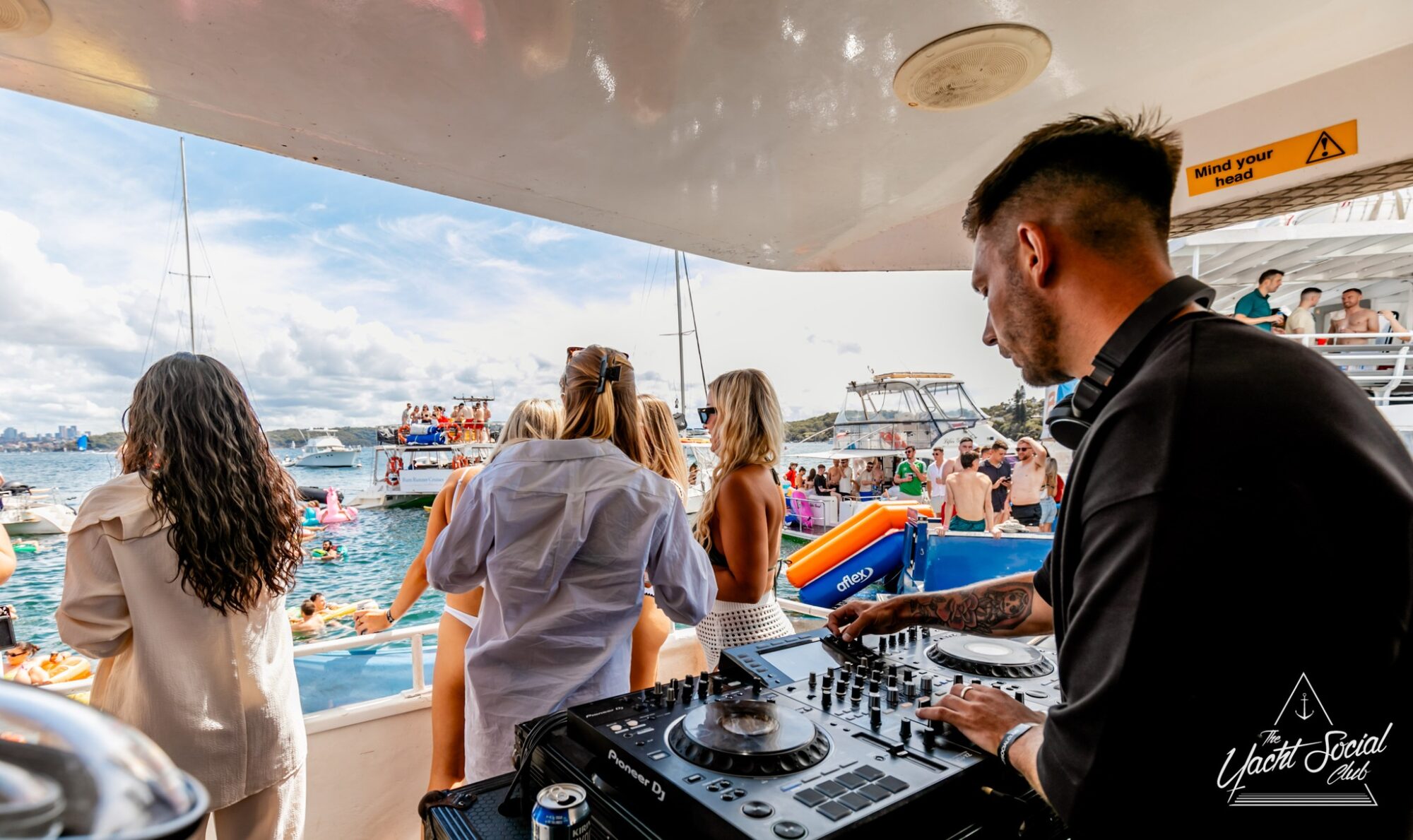 A DJ is spinning tracks on a boat with a turntable and mixer. People are dancing and enjoying the vibrant party on the deck, surrounded by other boats and a blue sky. It captures the lively atmosphere of Sydney Harbour Boat Hire The Yacht Social Club's outdoor event on the water.