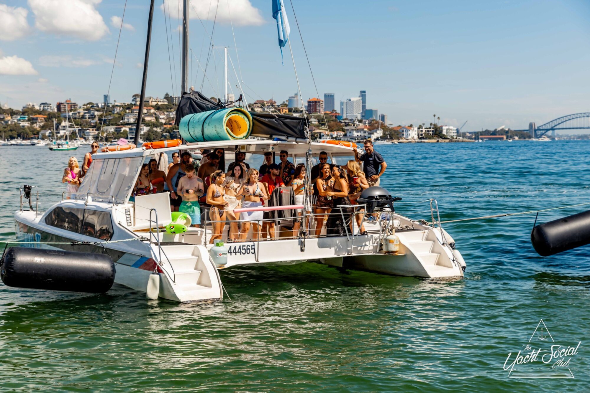 A group of people are gathered on a catamaran yacht, enjoying a sunny day on the water. The Yacht Social Club event is anchored near a city shoreline with tall buildings and a bridge visible in the background. The sky is clear with some scattered clouds.