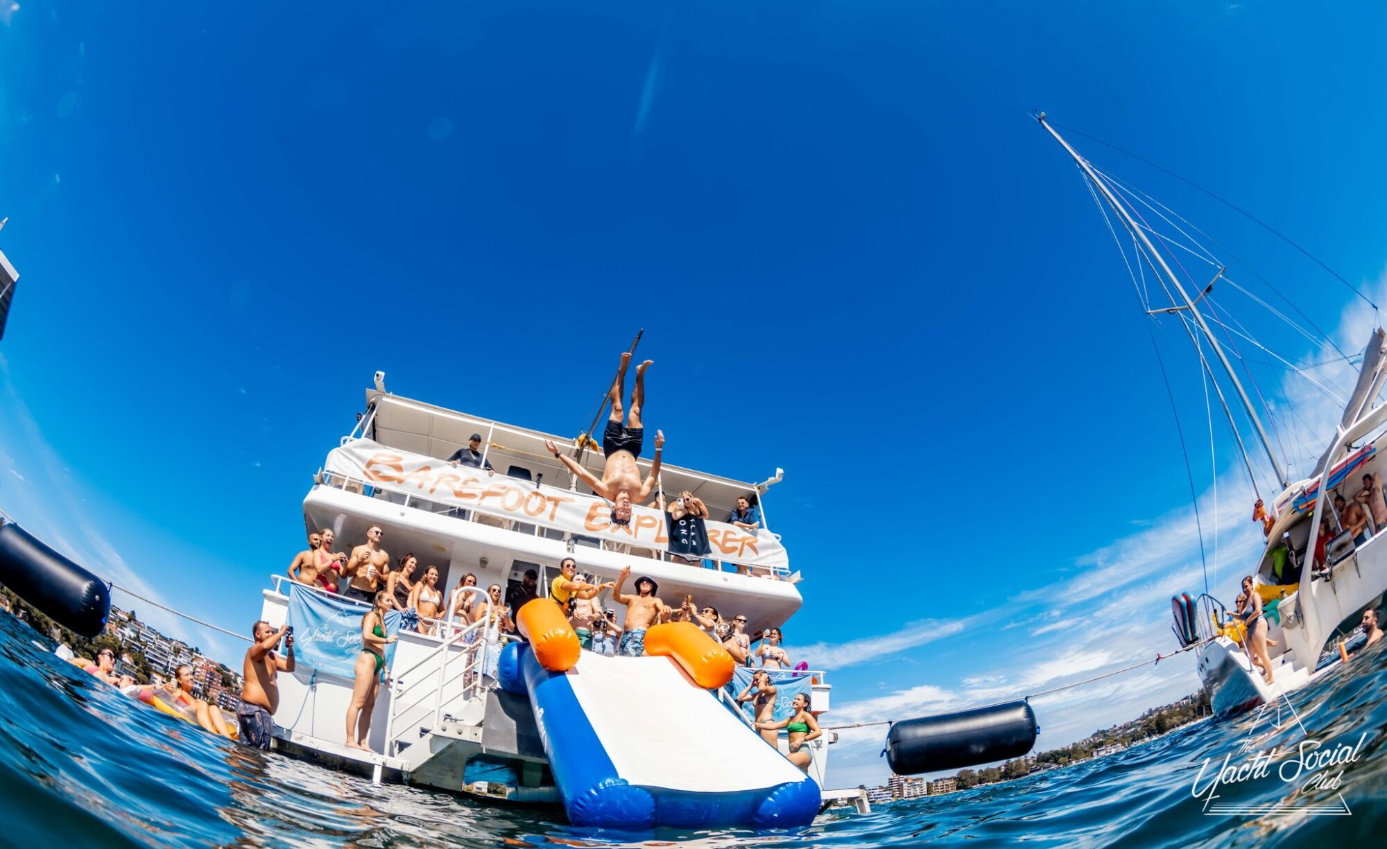 A group of people enjoying a day on "Self Explorer," an extravagant vessel from Sydney Harbour Boat Hire The Yacht Social Club, featuring an inflatable slide. Some are on the upper deck, others gather around the slide or swim in the clear blue waters, with one person mid-dive from above.