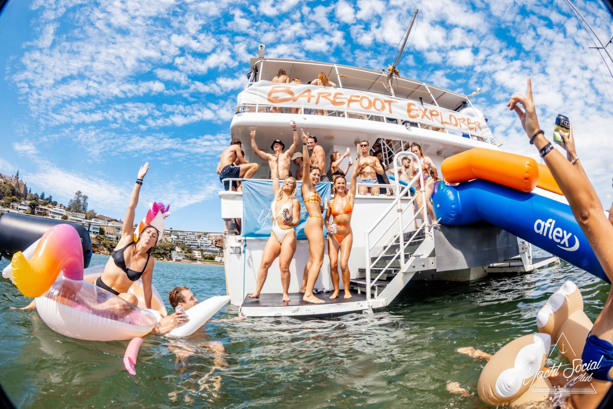 A group of people in swimwear having fun on a boat labeled "Barefoot Explorer." Some are dancing on the deck while others are in the water on floaties, including a unicorn-shaped one. The weather is sunny with a partly cloudy sky in the background, setting the perfect scene for Boat Parties Sydney with The Yacht Social Club.