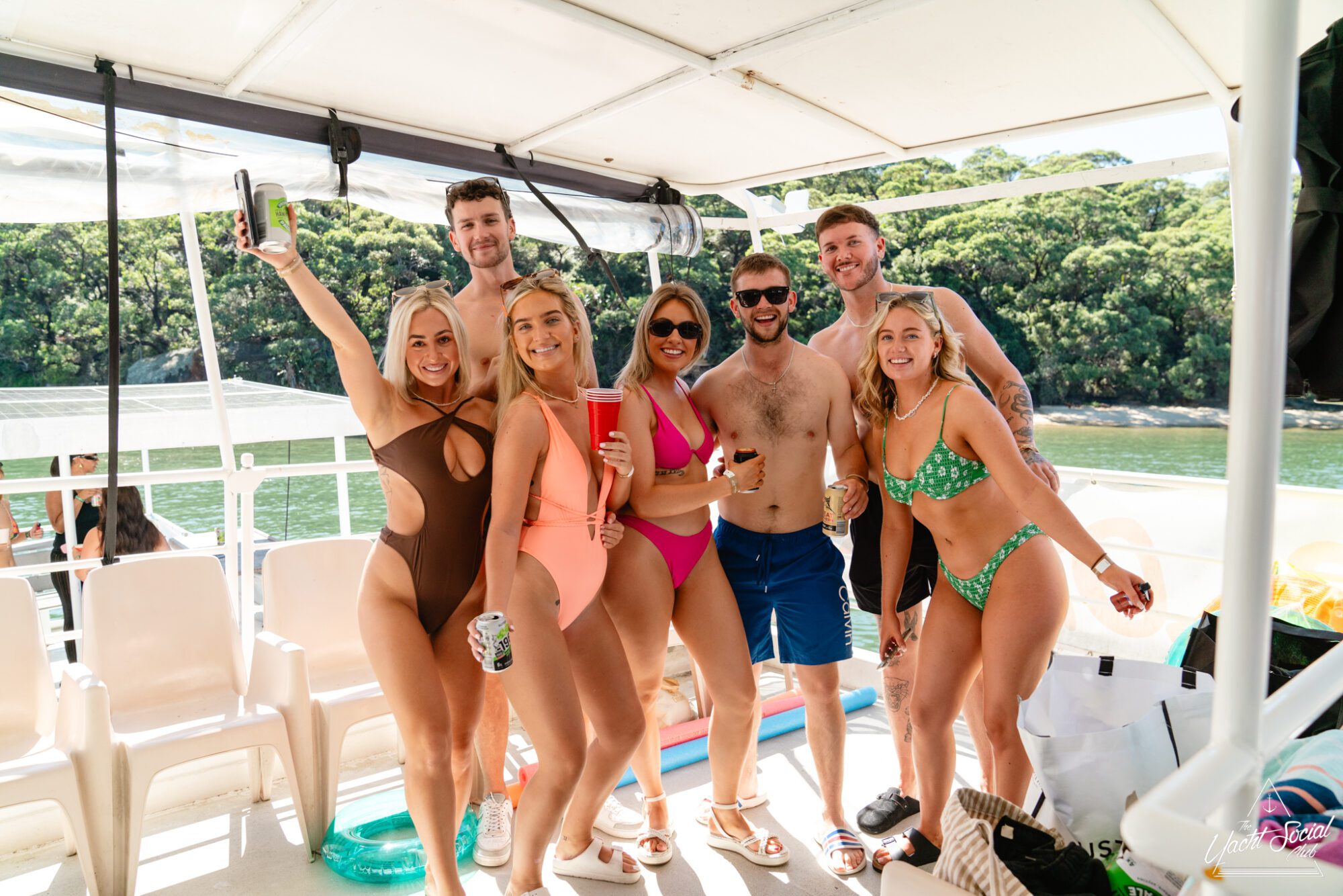 A group of seven people, dressed in swimsuits, pose happily on a boat from The Yacht Social Club Sydney Boat Hire. They are smiling and holding drinks. The boat is docked near a lush, green shoreline. The atmosphere appears festive and relaxed, with sunny weather enhancing the scene.