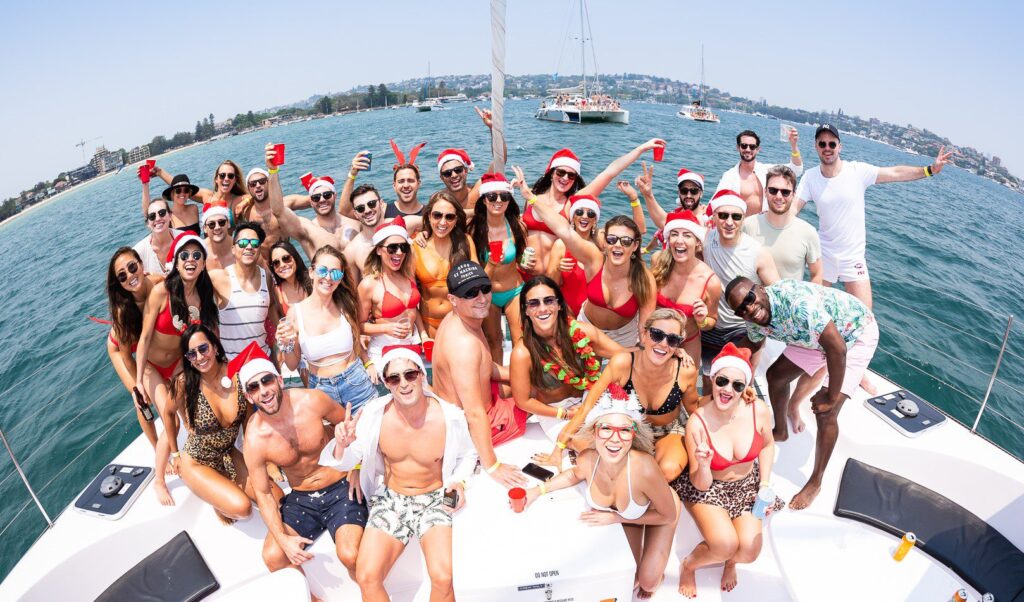 A large group of people partying on a boat, wearing swimsuits and Santa hats. They are smiling, cheering, and holding drinks, with a scenic waterfront and sailboats in the background. The atmosphere is lively and festive. Experience similar fun with Sydney Harbour Boat Hire at The Yacht Social Club.