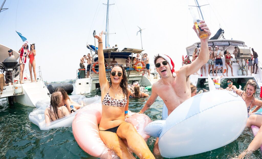 A lively scene of The Yacht Social Club Sydney Boat Hire, with numerous people enjoying themselves on the water. Two individuals are prominently featured in the front, smiling and holding drinks while floating in inflatable rings. Several boats and more partygoers are visible in the background.