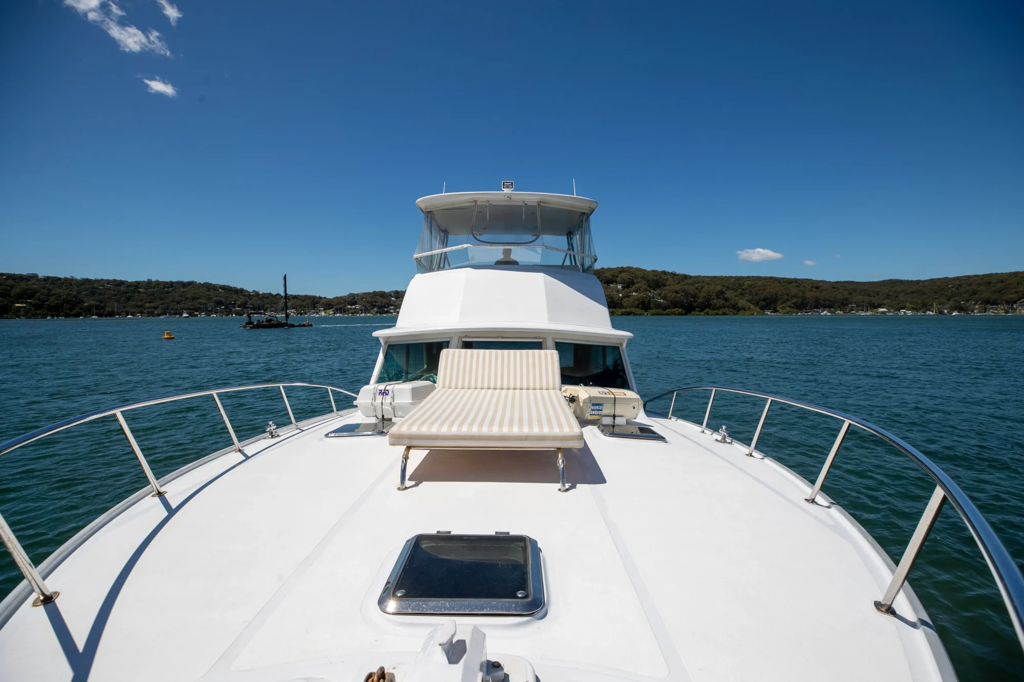 A view of the deck of a white yacht sailing on calm blue waters under a clear sky. The deck features a padded sun lounger, rails, and a clear glass hatch. Distant green hills and a few boats can be seen near the shoreline in the background, perfect for a Luxury Yacht Rentals Sydney experience.