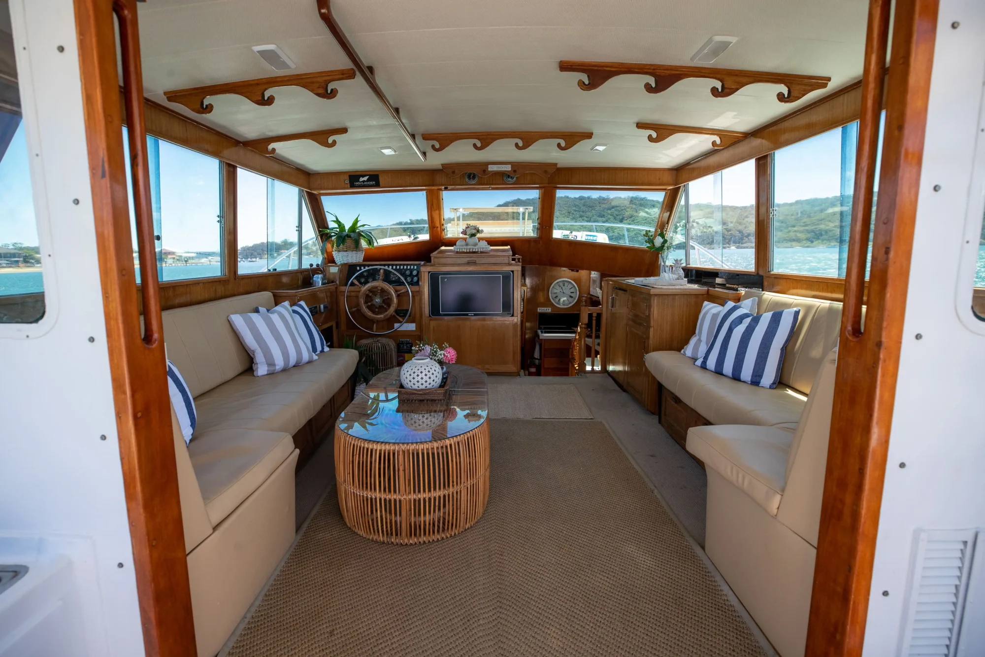 Interior view of a beautifully furnished boat cabin, featuring two beige sofas with striped cushions, a wicker coffee table with decor, and a steering wheel at the front. Large windows offer scenic views of water and distant greenery, filled with natural light—perfect for The Yacht Social Club Sydney Boat Hire experience.