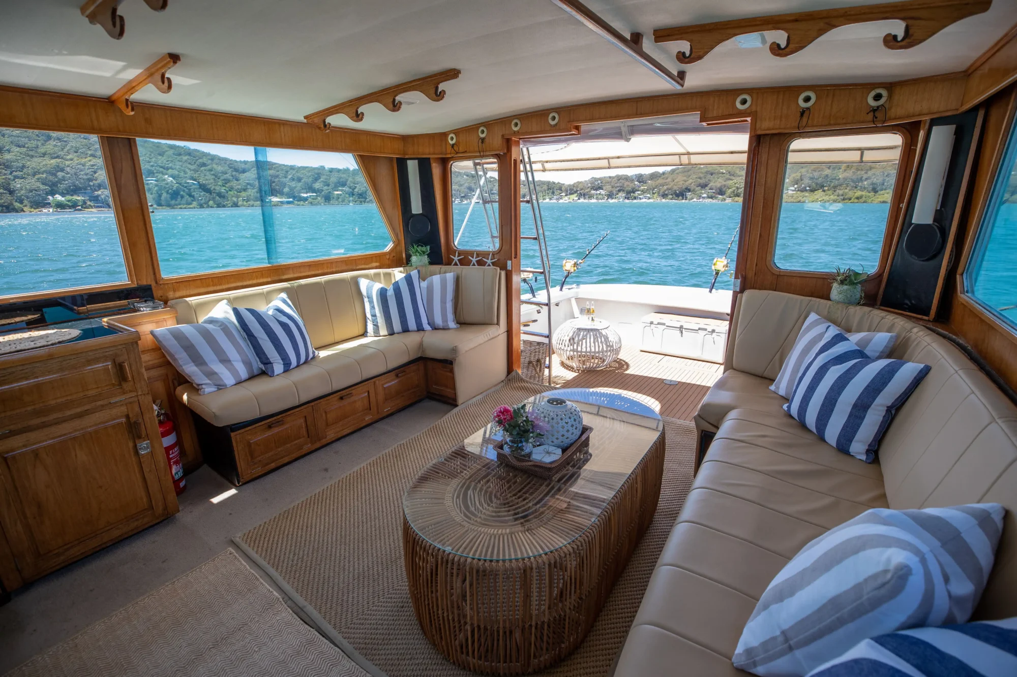 Interior of a boat with large windows offering views of a scenic waterway. There are two L-shaped beige leather sofas with striped pillows, a wooden coffee table, and a neutral rug. Plants and decorative items accent the cozy, well-lit space—perfect for The Yacht Social Club Sydney Boat Hire or Luxury Yacht Rentals Sydney.