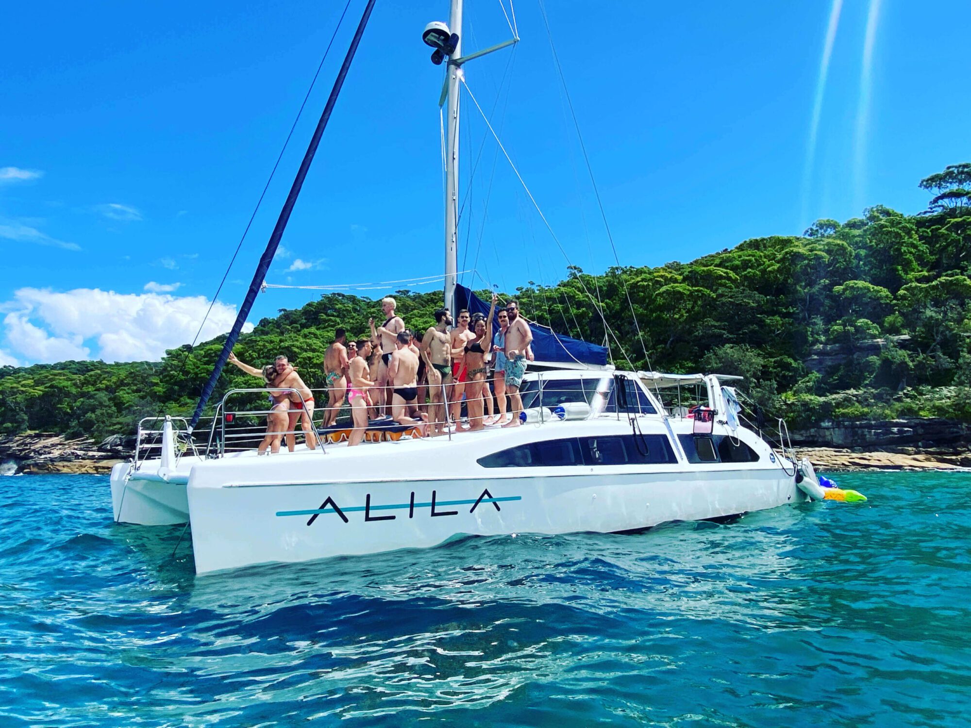 A group of people stand on a white catamaran sailboat named "ALILA." The boat is anchored in clear, turquoise water near a lush, green shoreline under a vibrant blue sky. Everyone appears to be enjoying a sunny day with The Yacht Social Club Sydney Boat Hire.