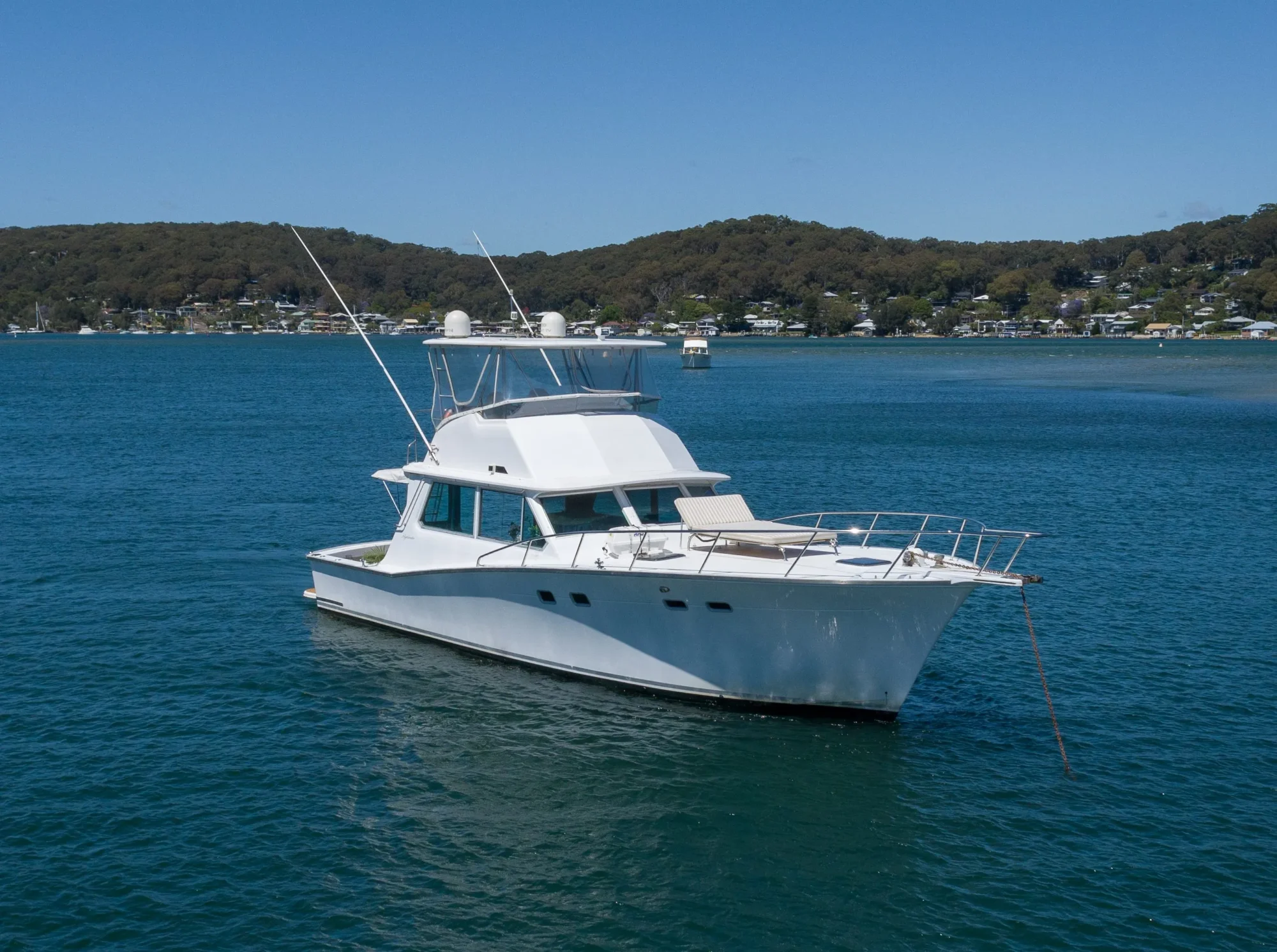 A medium-sized white motor yacht anchored on a calm blue body of water with a tree-lined shoreline and small houses visible in the background under a clear blue sky. Perfect for a day out with The Yacht Social Club Sydney Boat Hire.