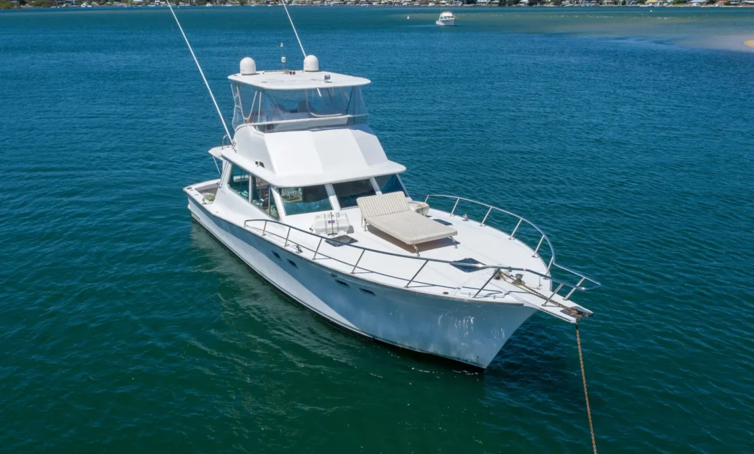 A white yacht anchored in calm blue-green waters near a shoreline with wooded hills and scattered houses in the background. The yacht, perfect for Boat Parties Sydney The Yacht Social Club, features a sun deck on the bow and an enclosed flybridge, with two antennas extending upwards.