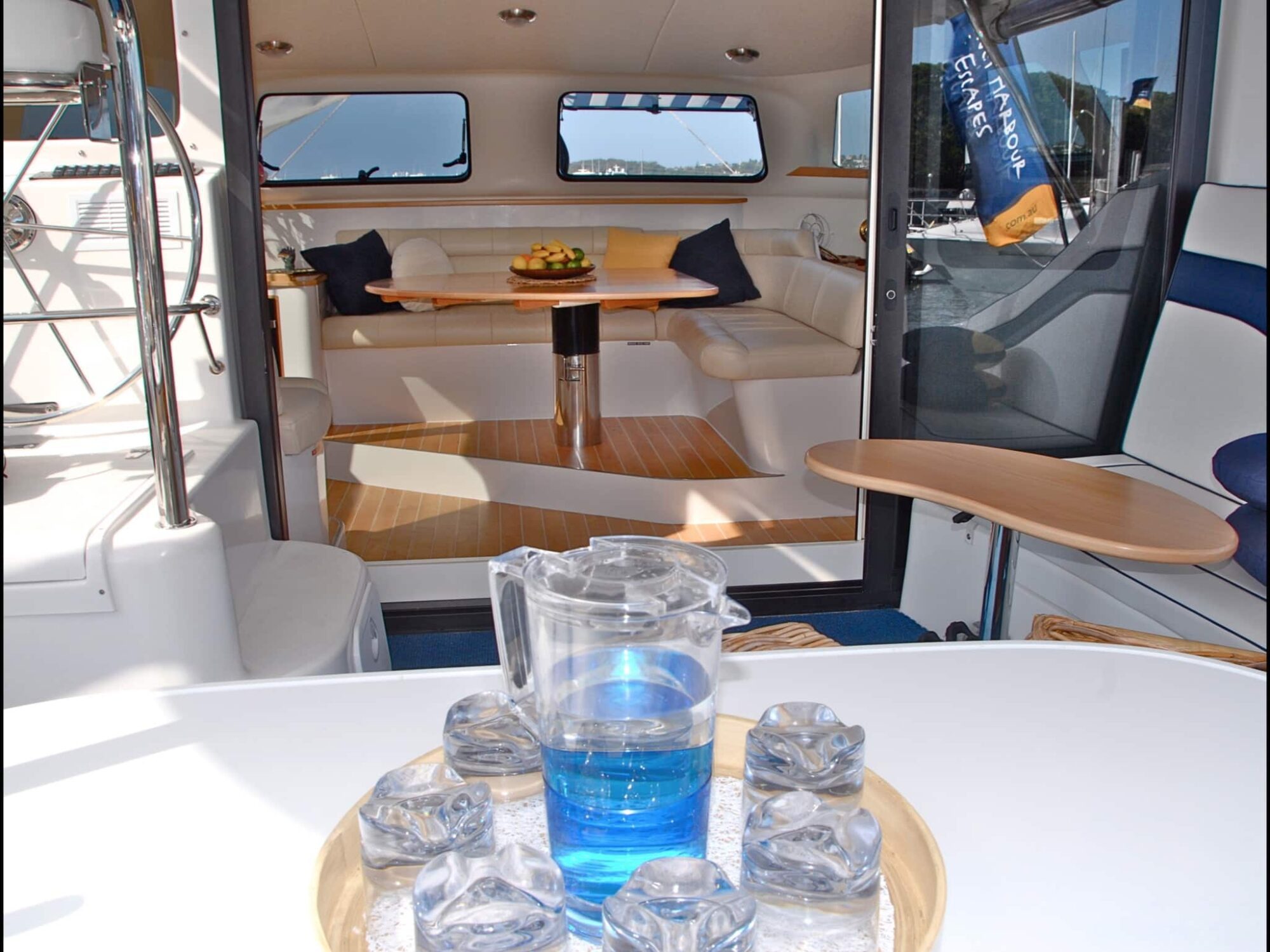 Interior view of a boat, featuring a cozy seating area with a round table and cushioned seats in the background. In the foreground, a glass pitcher filled with blue water and six glasses with ice cubes are arranged on a round tray. A flag is visible through the window, perfect for The Yacht Social Club events.