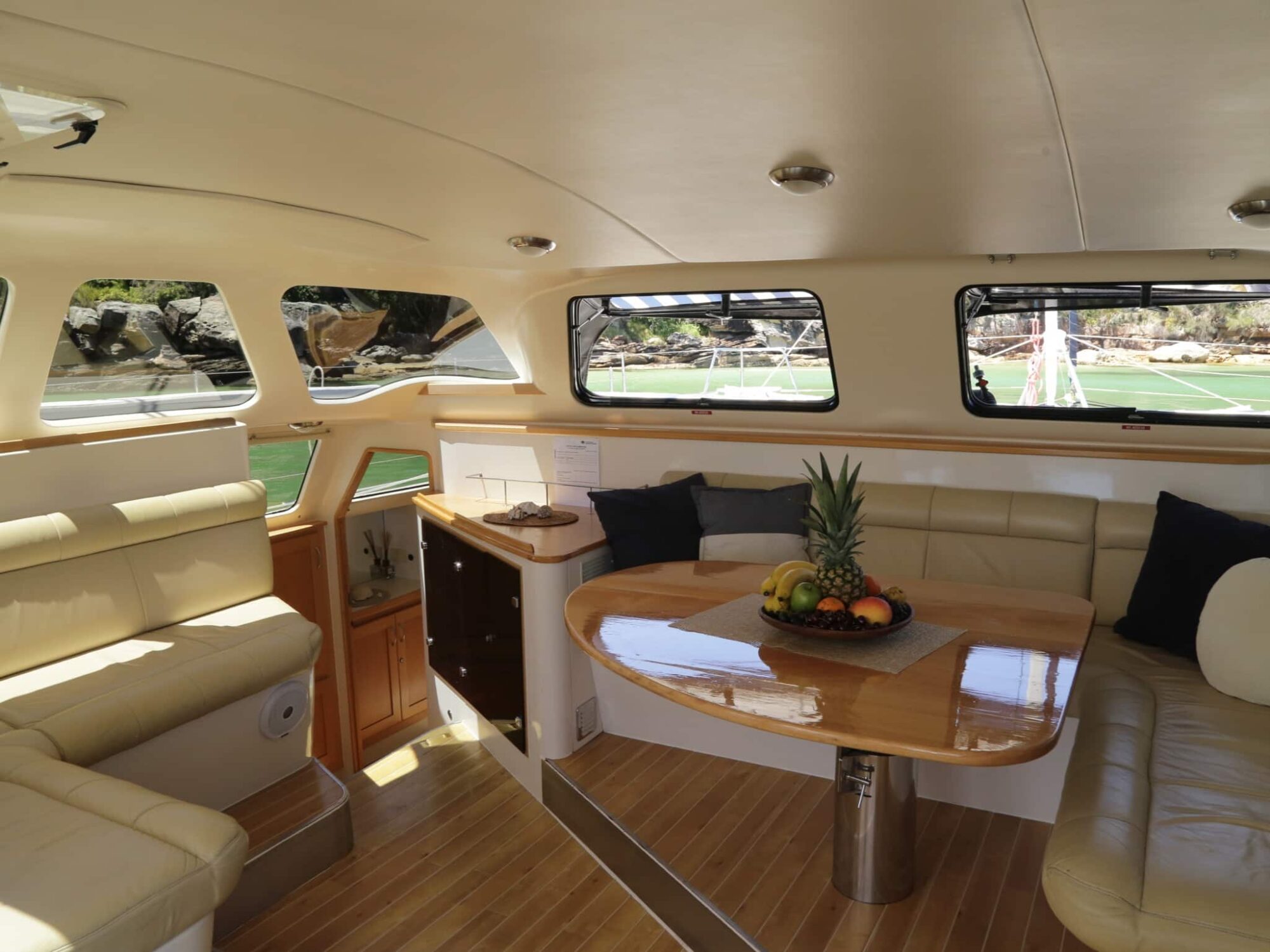 The interior of a yacht features an upholstery seating area with cushions around a polished wooden table. The table is adorned with a fruit basket centerpiece. Large windows surround the space, providing ample natural light and views of an outdoor deck—perfect for Boat Parties Sydney The Yacht Social Club.