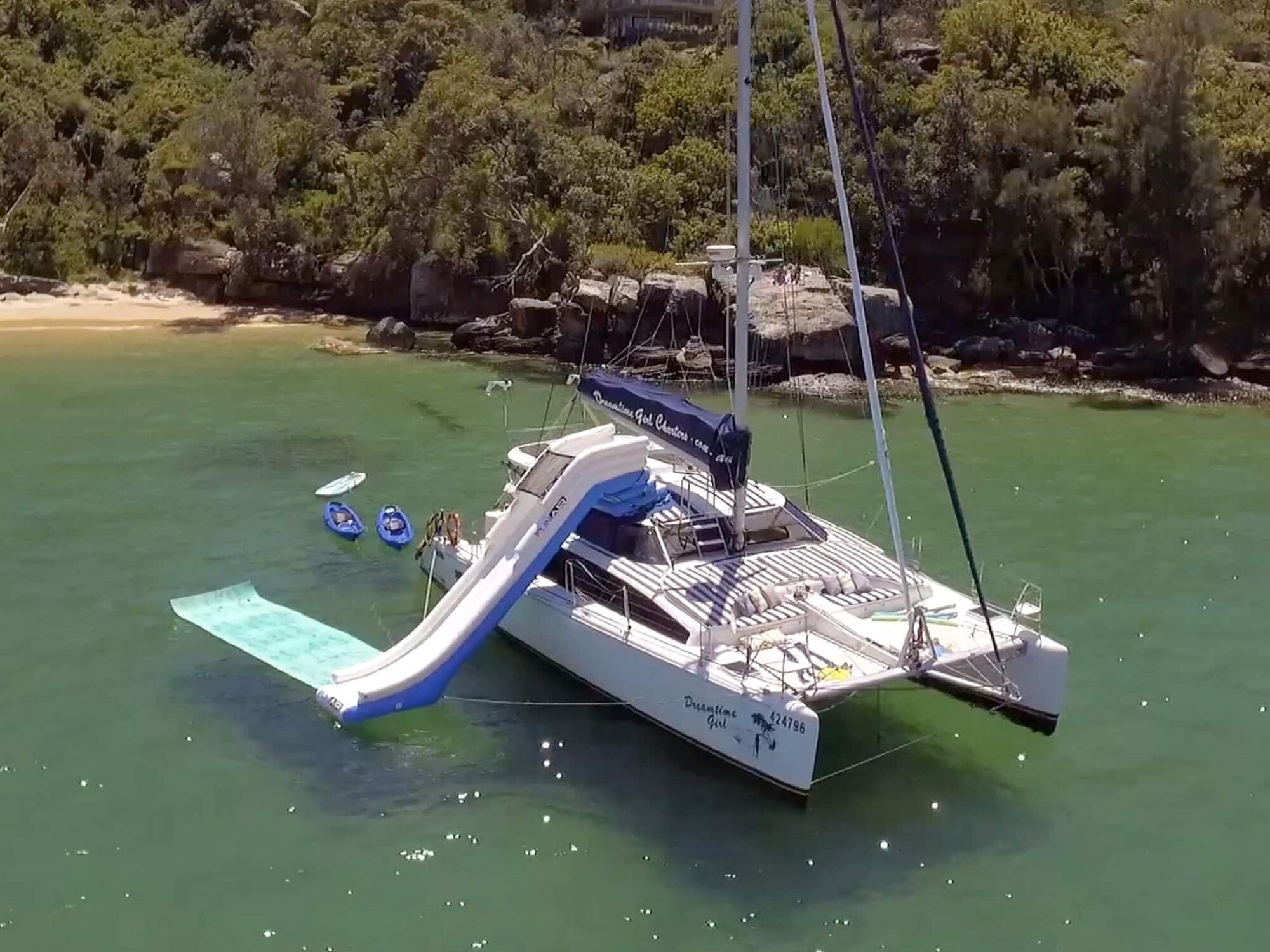 A catamaran is anchored near a leafy shoreline, featuring a blue slide extending into the water and a floating mat beside it. Paddleboards and other water toys are visible, with people enjoying the activities around The Yacht Social Club Sydney Boat Hire boat.