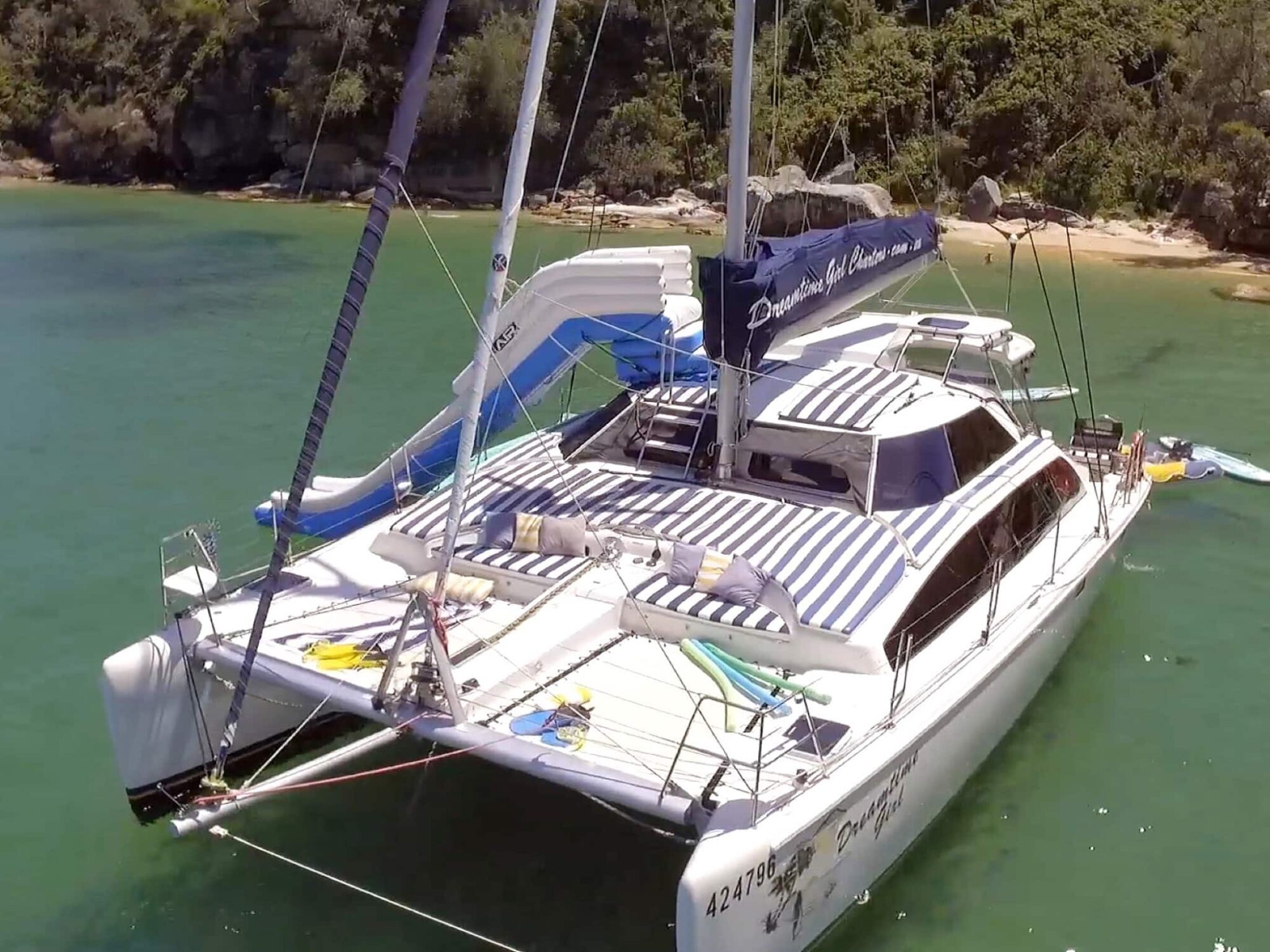 A white catamaran with a striped sunshade and blue accents is anchored in a turquoise bay near a verdant shore. The boat, available through Luxury Yacht Rentals Sydney, has a slide extending from its deck into the water. The deck is equipped with padded seating and various recreational items.