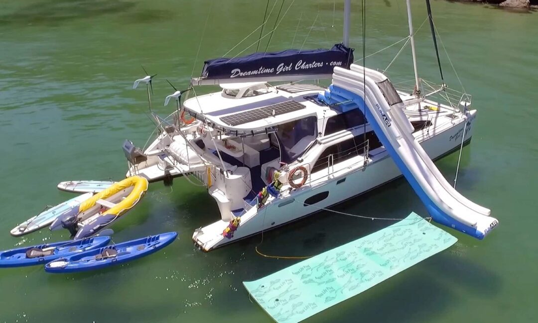 A catamaran anchored in clear green water is equipped with kayaks, a water slide, and a floating mat. The boat has a shaded deck, solar panels, and the banner "Dreamtime Girl Charters" for Boat Rental and Parties Sydney. The shoreline with rocks and trees is visible in the background.