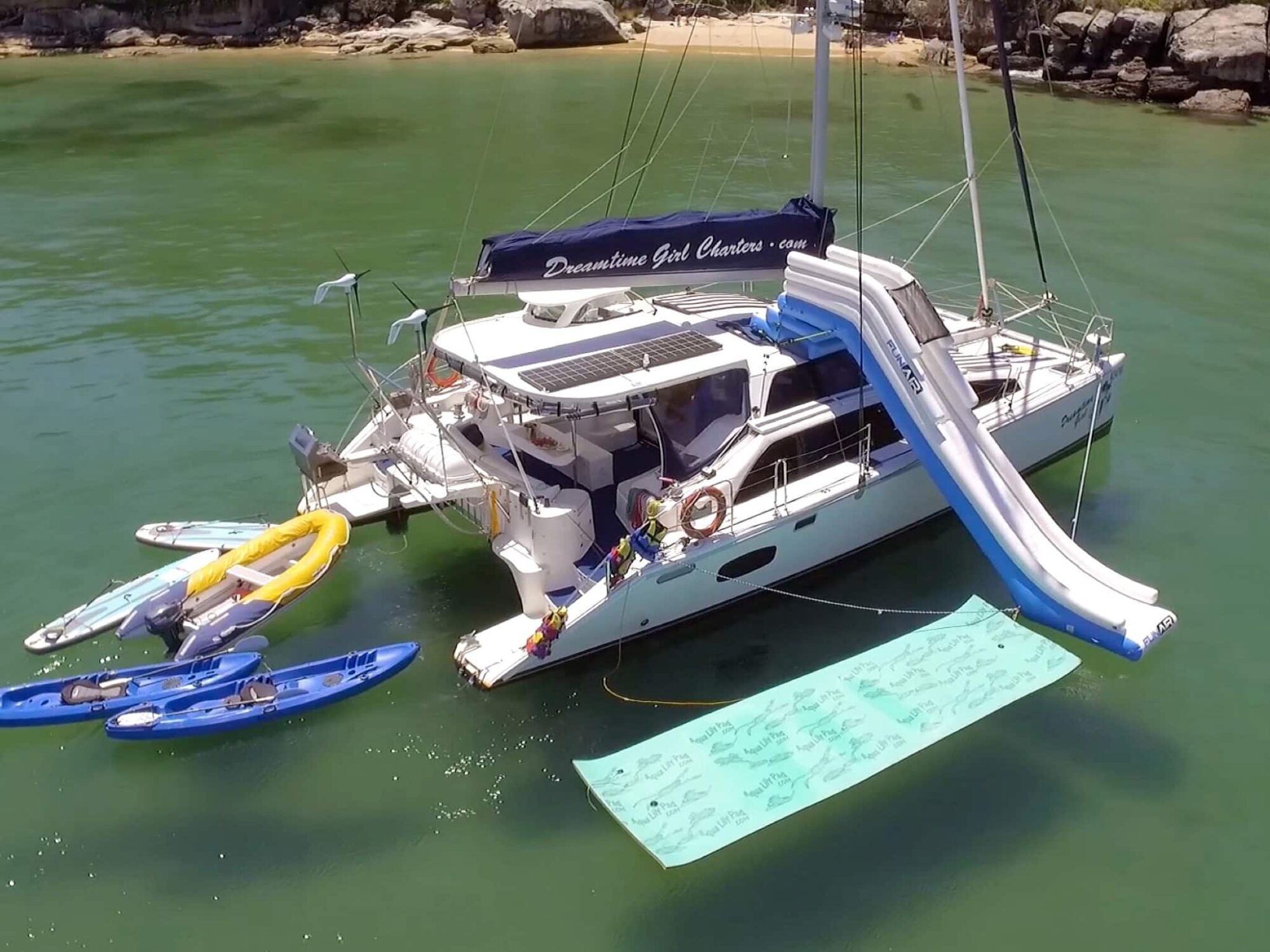 A catamaran anchored in clear green water is equipped with kayaks, a water slide, and a floating mat. The boat has a shaded deck, solar panels, and the banner "Dreamtime Girl Charters" for Boat Rental and Parties Sydney. The shoreline with rocks and trees is visible in the background.