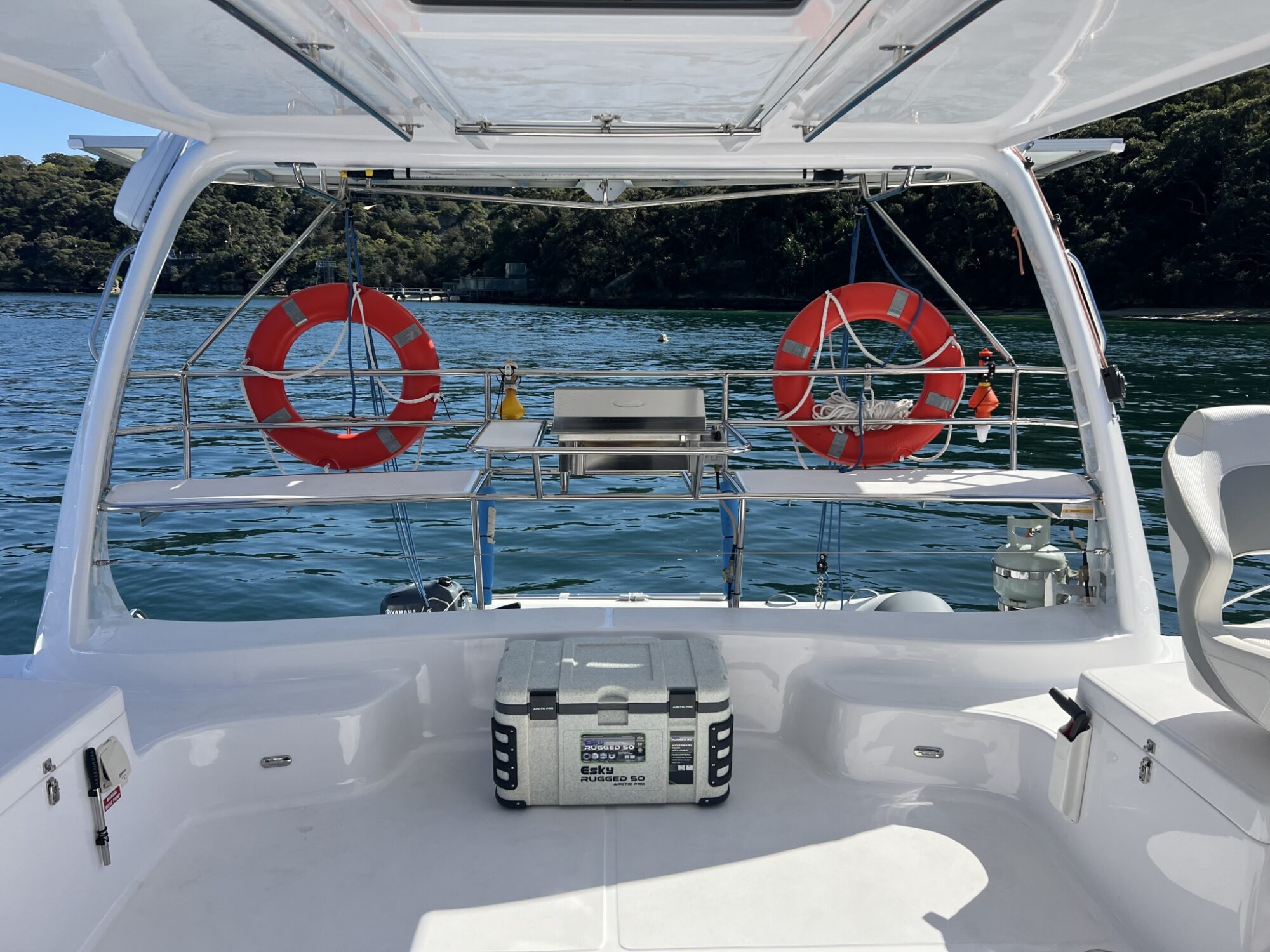 A white boat deck equipped with storage compartments, two life rings attached to the railing, and a cooler at the center. The boat is docked on a calm blue body of water with forested land visible in the background under a clear sky. Ideal for Luxury Yacht Rentals Sydney and Boat Parties Sydney The Yacht Social Club.