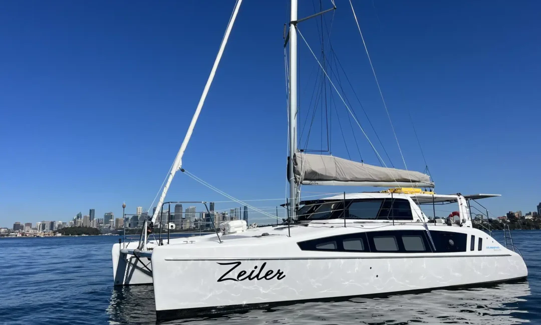 A white catamaran sailboat named "Zeiler" glides on calm, clear blue waters with a city skyline in the background under a cloudless sky. The boat features a modern design with large windows and a grey-sailed mast, perfect for Luxury Yacht Rentals Sydney from The Yacht Social Club.