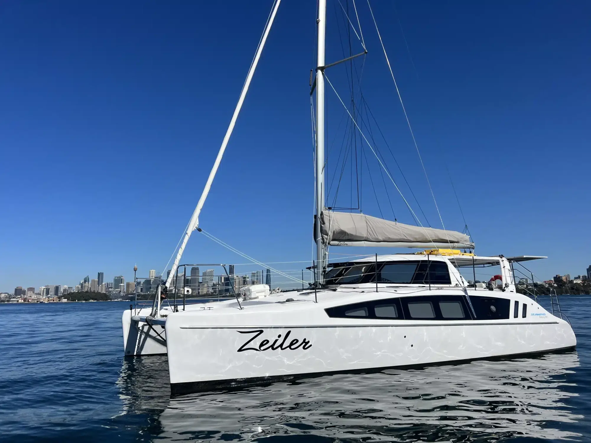 A white catamaran sailboat named "Zeiler" glides on calm, clear blue waters with a city skyline in the background under a cloudless sky. The boat features a modern design with large windows and a grey-sailed mast, perfect for Luxury Yacht Rentals Sydney from The Yacht Social Club.