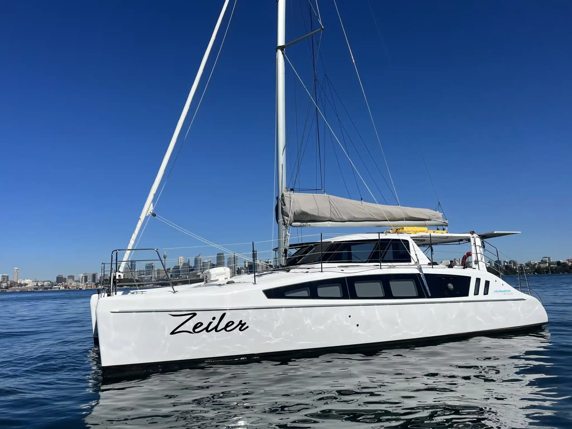 A sleek white sailing catamaran named "Zeiler" is anchored on calm waters with a cityscape visible in the background under a clear blue sky. Perfect for Boat Parties Sydney The Yacht Social Club, it features a large mainsail rolled up on the boom and several cabins with windows.