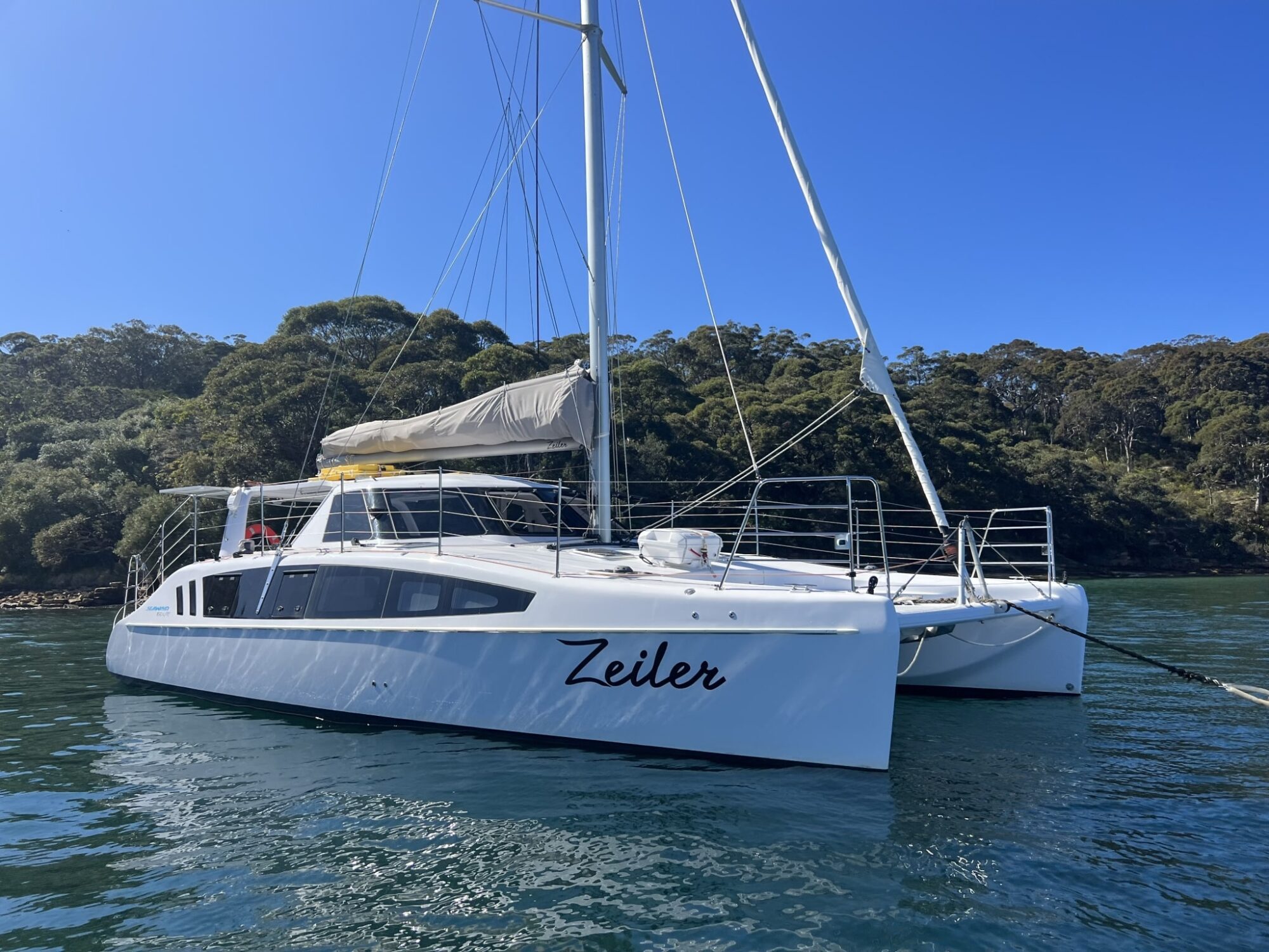 A white catamaran named "Zeiler" is anchored in calm, clear blue water. The boat has a sleek design with large windows on the sides and a sail furled on its main mast, perfect for Boat Parties Sydney The Yacht Social Club. Lush green trees line the background under a sunny, clear blue sky.