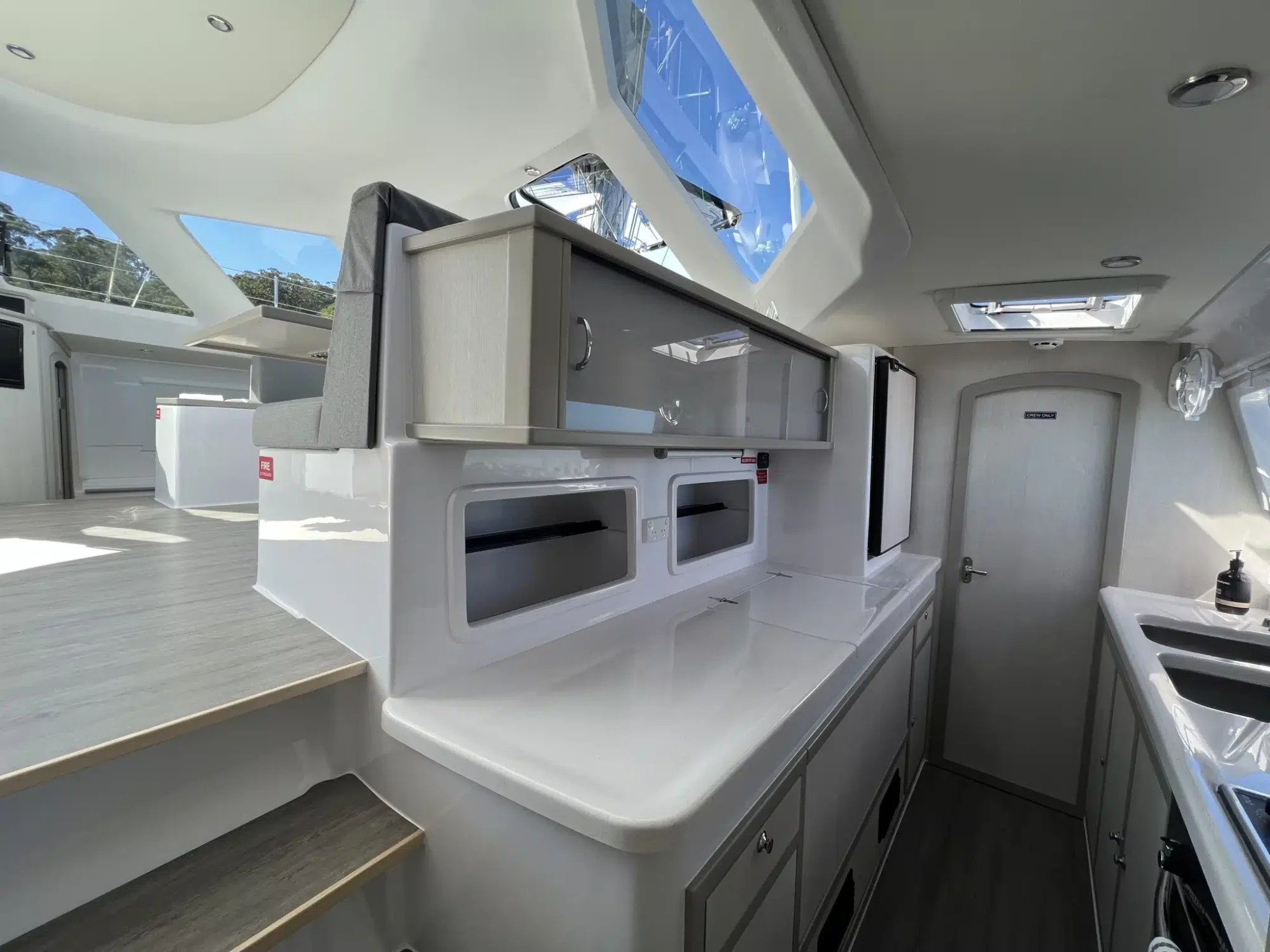 A modern yacht interior featuring a sleek kitchen with white countertops, overhead cabinets, and stainless steel appliances. Two steps lead up to a cozy seating area with large windows providing ample natural light. Perfect for Luxury Yacht Rentals Sydney. A door at the back opens to another section of the yacht.