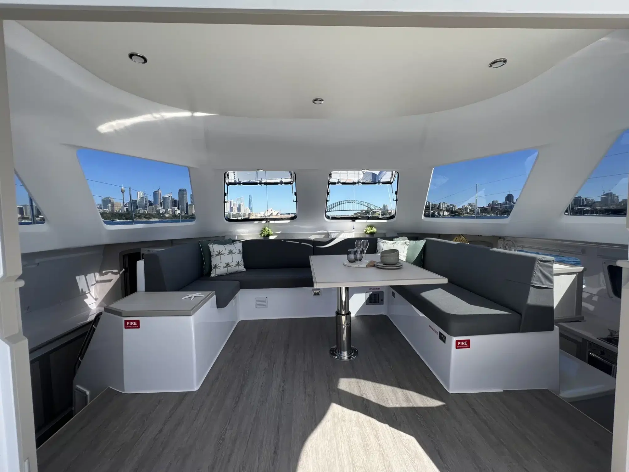 A modern yacht interior featuring a spacious seating area with gray cushions and a white, adjustable table at the center. Large windows surround the space, providing panoramic views of a city skyline and bridge outside. The Yacht Social Club offers luxury yacht rentals in Sydney for unforgettable experiences.