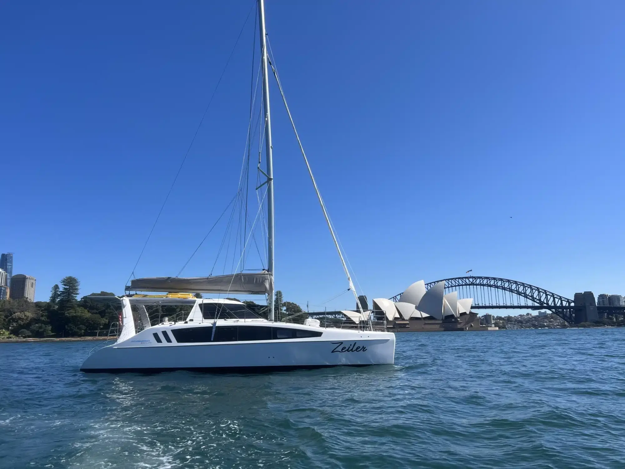 A catamaran with the name "Zeilco" sails on a calm body of water. In the background, Sydney Opera House and Sydney Harbour Bridge are visible under a clear blue sky. Experience this unique view with The Yacht Social Club's Sydney Harbour Boat Hire for an unforgettable adventure.