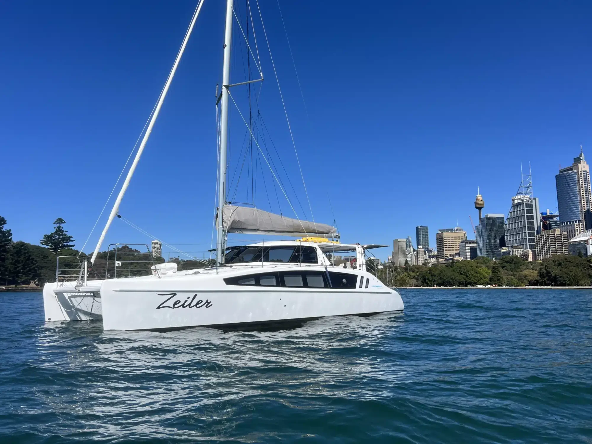 A white sailboat named "Zeiler" is anchored on water with a clear blue sky above. City skyscrapers, greenery, and the iconic Sydney Tower are visible in the background, perfect for enjoying Boat Parties Sydney The Yacht Social Club.