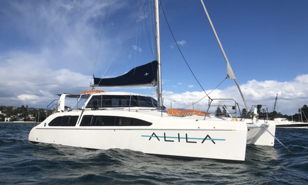 A white catamaran named "ALILA" is anchored in calm, dark blue waters under a partly cloudy sky. Featuring a blue sail and several masts, the scene is complemented by houses and greenery in the background. Perfect for Boat Parties Sydney The Yacht Social Club or Luxury Yacht Rentals Sydney experiences.