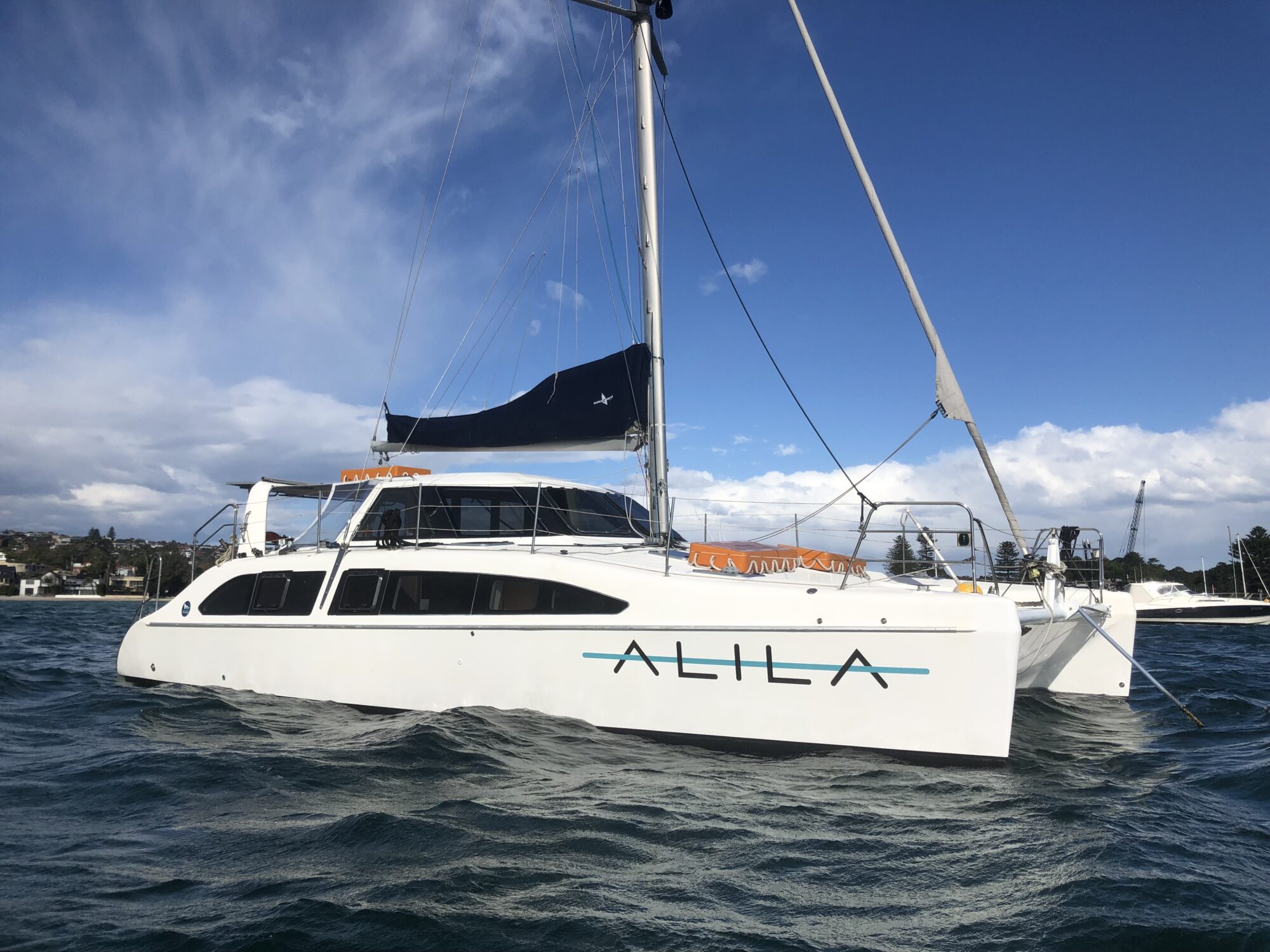 A white catamaran named "ALILA" is anchored in calm, dark blue waters under a partly cloudy sky. Featuring a blue sail and several masts, the scene is complemented by houses and greenery in the background. Perfect for Boat Parties Sydney The Yacht Social Club or Luxury Yacht Rentals Sydney experiences.