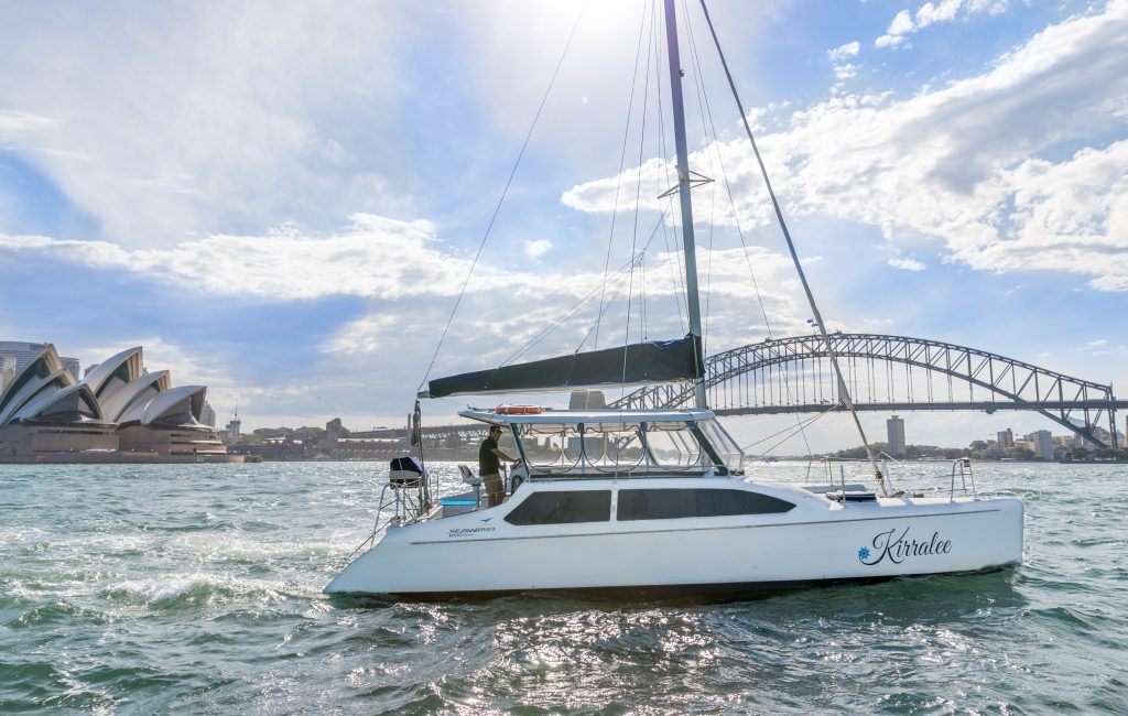 A sailboat named "Kiralee" navigates through water with the iconic Sydney Opera House and Sydney Harbour Bridge visible in the background. The sun shines brightly, illuminating a clear day with a few scattered clouds, perfect for a luxurious experience like those with Luxury Yacht Rentals Sydney.