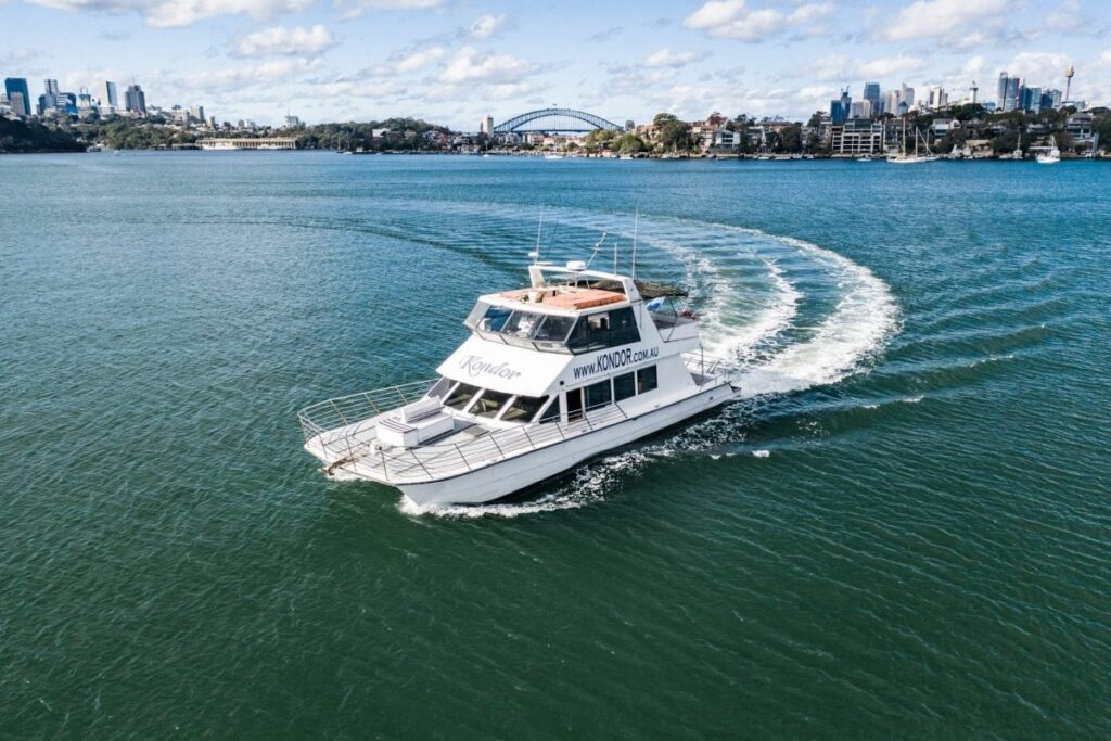 A white boat named "Kondor" from Sydney Harbour Boat Hire by The Yacht Social Club sails through calm, blue waters, leaving a distinct wake behind. The city skyline and a landmark bridge are visible in the background under a partly cloudy sky.